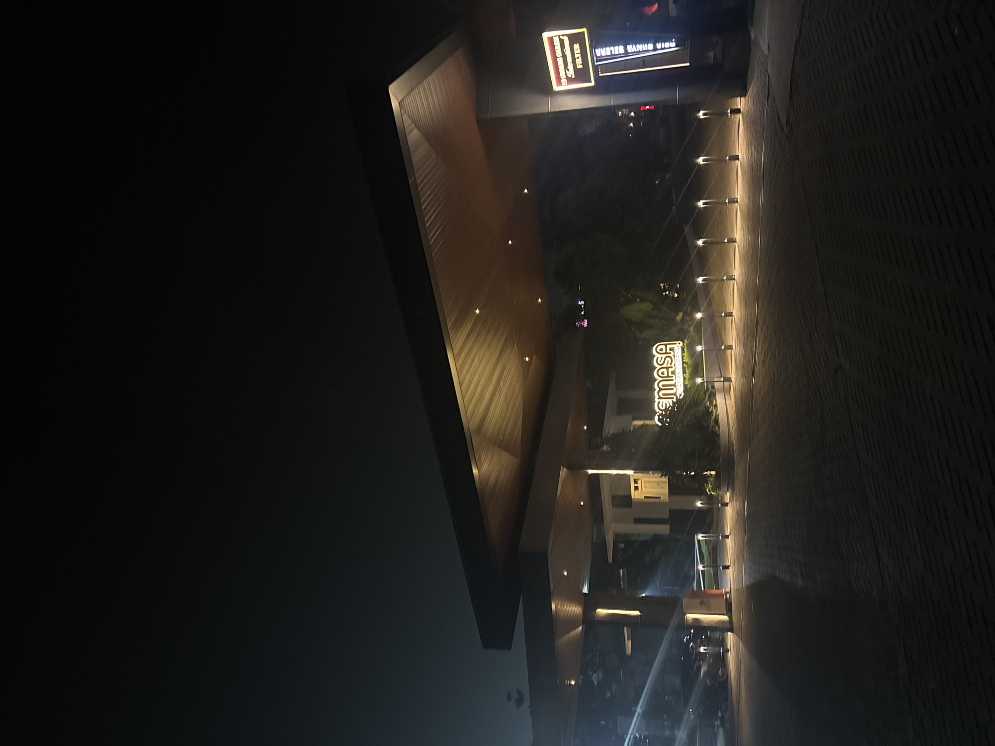 The image shows a nighttime scene outside a building with modern architecture. The building features a wooden-paneled overhang illuminated by embedded lights. In the background, there are illuminated signs. One sign, which appears to be a neon sign, reads "SEMASA" in large, stylized letters. Another sign above it, also illuminated, has text that is partially legible, including "GARAGE" and "FILTER" in smaller print on a dark background with a red border. The ground is paved with brick-like stones, and a row of evenly spaced bollards with upward-facing lights lines a walkway. The overall ambiance suggests a well-lit commercial or public space at night, possibly an entrance to a shopping center or entertainment venue, given the prominent signage. The location is identified as Babakan Madang, Indonesia. There are no people clearly visible in the image. The lighting suggests it is evening or nighttime.