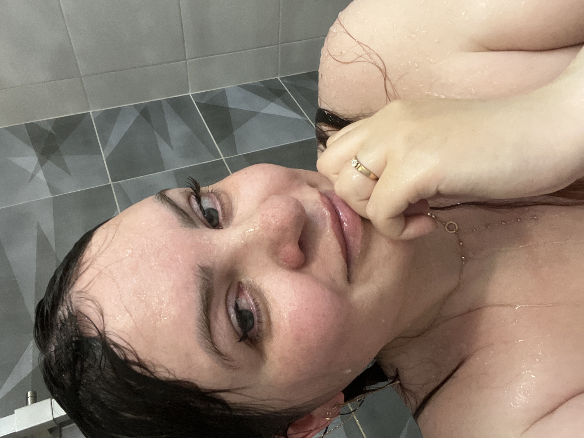 A close-up, rotated selfie photograph shows a wet, fair-skinned individual with dark hair and eyes. The person is holding their right hand near their face, with a gold ring visible on their finger. A gold necklace is also visible around the person's neck. Water droplets are present on the skin and hair, suggesting recent exposure to water. The background consists of a gray tiled wall and floor. The location is not discernible from the provided image.