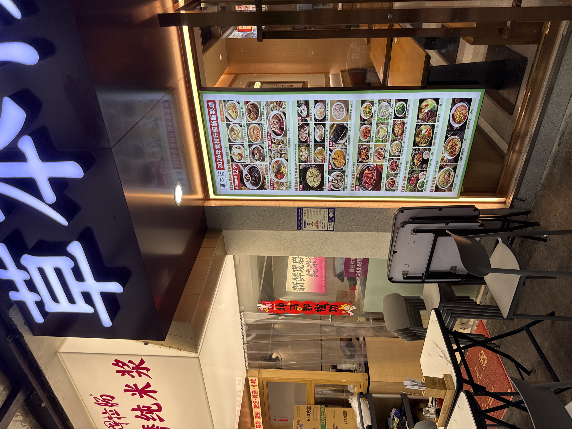 The image shows a restaurant storefront in Haizhu District, China. The restaurant's name, "草本汤" (Cao Ben Tang), is displayed in large, illuminated characters above the entrance. A menu board is displayed prominently in the window, showcasing various dishes with accompanying prices. Outside the restaurant, several tables and chairs are set up for outdoor dining. A red and white sign with Chinese characters hangs vertically on the window, possibly indicating a promotion or special. The storefront appears to be in an urban setting, with a dimly lit background suggesting it is evening. Boxes are stacked near the entrance, and some kitchen equipment is visible through the window. The overall scene depicts a typical restaurant environment in a bustling city.