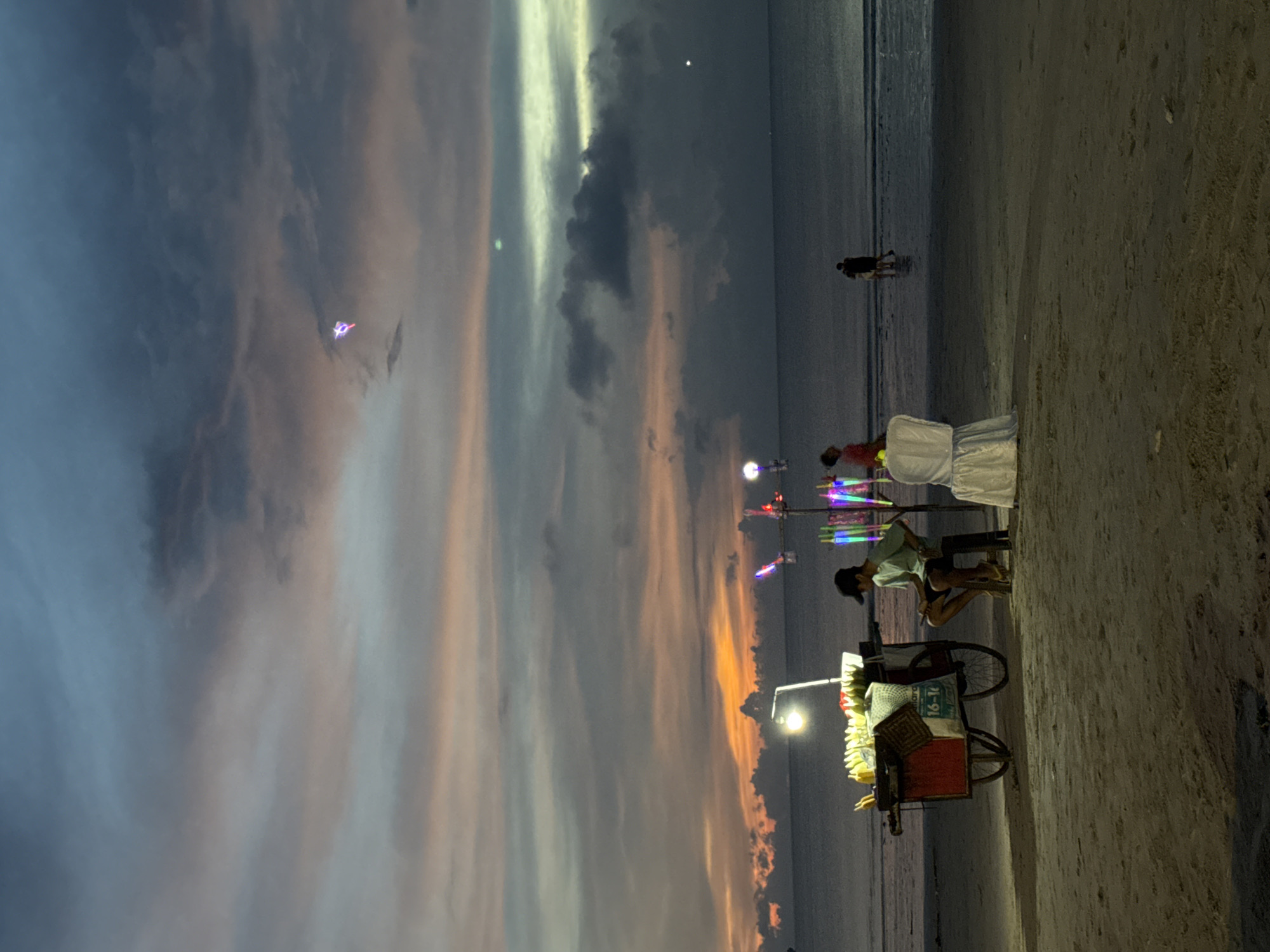The image shows a beach scene at dusk, likely in Jimbaran, Indonesia. A street vendor is visible with a cart selling grilled corn, illuminated by a light. People stand near the cart, possibly customers. Further down the beach, other individuals are seen walking. The sky is filled with clouds, and an orange glow is present at the horizon.