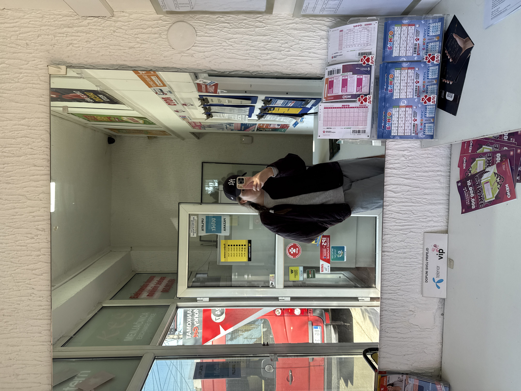 The photograph captures a reflection in a mirror that shows a person taking a selfie inside what appears to be a small shop or kiosk. The person is holding a smartphone, visible in the mirror, and is wearing a cap and a dark jacket over a light-colored top. The reflection reveals the interior of the kiosk, showing a window and glass door where a red bus can be seen outside.
Various advertisements and lottery tickets are displayed on the wall above the counter. There are signs for lottery games and mobile services, specifically Telenor's VIP service. The glass door shows various stickers and advertisements.
The kiosk also features an "Exchange Office" sign, written as "MENJAČNICA," indicating it is a place where currency exchange is possible. The sign also says "UPLATA PAZARA." The street names are not visible, making it impossible to precisely locate the photo within the city of Vozdovac Urban Municipality, Serbia, or to confirm its location.