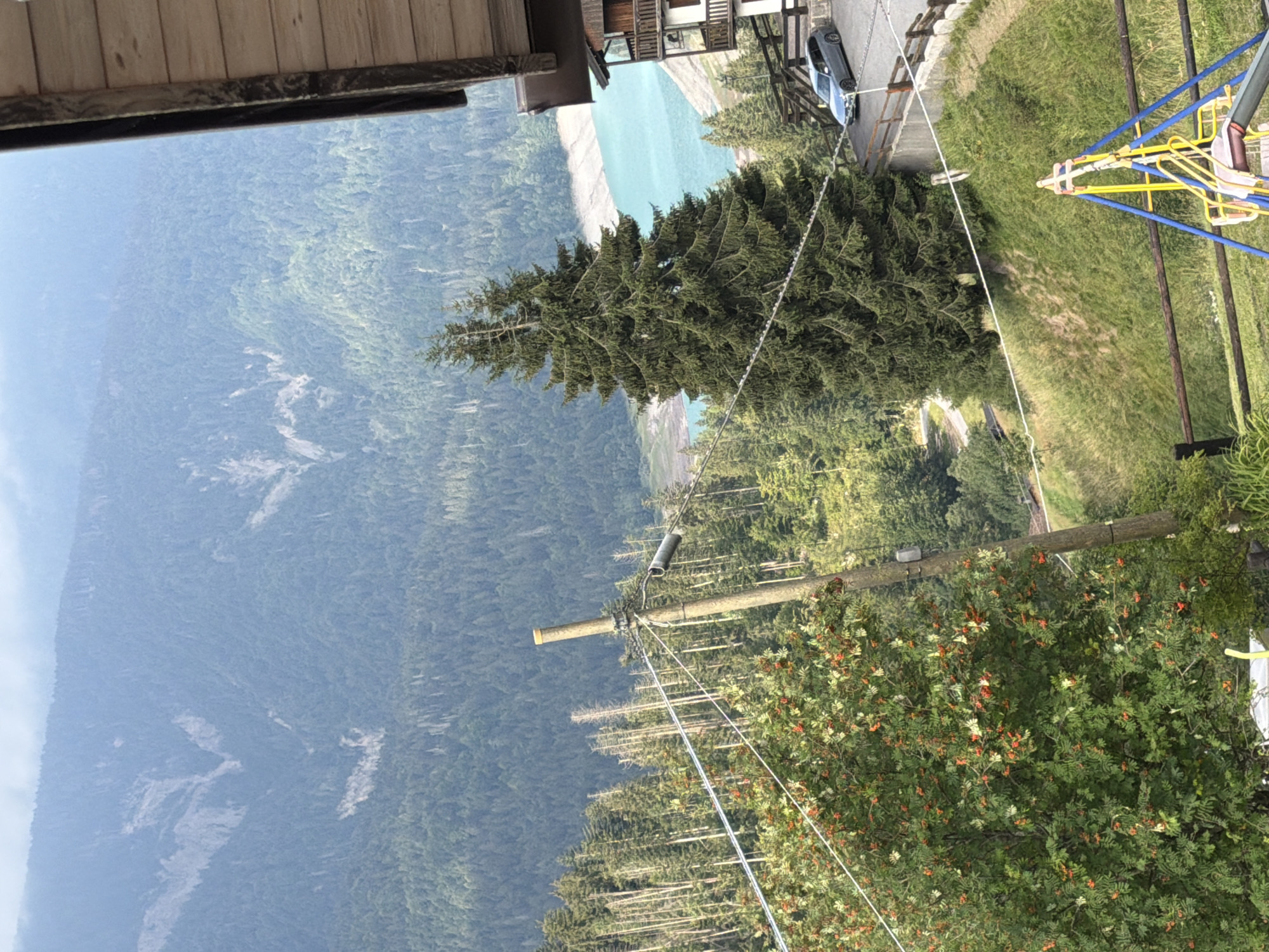This image depicts a mountain landscape with a body of turquoise water in the background, likely a lake. The foreground features a densely wooded slope with a building on the right side. A tall evergreen tree stands prominently in the middle ground. There are power lines running across the scene, supported by wooden poles. In the lower right corner, a colorful children's swing set is visible. A silver car is parked on a paved area next to the building. The scene is bathed in natural light, suggesting it was taken during the day. The presence of mountains, forests, and a lake strongly suggests a location in the Italian Alps, possibly within the region of Sauris / Zahre, Italy, known for its natural beauty and mountainous terrain.