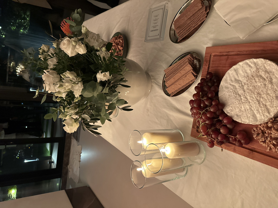 The scene is set indoors at night within a private residence or event space located in London, United Kingdom. A long table, covered with a white tablecloth, is arranged with food and decorative elements.
In the left foreground, three lit pillar candles of varying heights are contained within clear glass hurricane lanterns. To their right, a rectangular wooden cutting board displays a cluster of red grapes, a round wheel of soft white cheese with a textured rind, and a small pile of walnuts. Further right, two oval silver platters hold stacks of flat, rectangular crackers or crispbreads.
Central to the table arrangement is a white ceramic vase filled with a substantial floral display. This arrangement primarily features white roses and white lisianthus, interspersed with green eucalyptus leaves and darker green foliage. A single reddish-orange protea-type flower is visible on the right side of the arrangement. Behind the flowers and to the right, a small, shallow grey-green bowl contains a quantity of light-colored snacks, possibly pretzels. Adjacent to this bowl, a clear acrylic stand holds a small white card with text, appearing to be a menu. A stack of folded white napkins is partially visible on the far right edge of the table.
The background reveals large glass doors with black frames, opening to a dark outdoor area. Several small, upright ground lights are visible beyond the doors, illuminating portions of the exterior. Faint human figures are discernible standing outside in the darkness. A white object, possibly a cloth, rests on the ground directly outside the glass doors. Indoors, on the far right, the lower leg of a person wearing dark trousers is partially visible standing near the table.