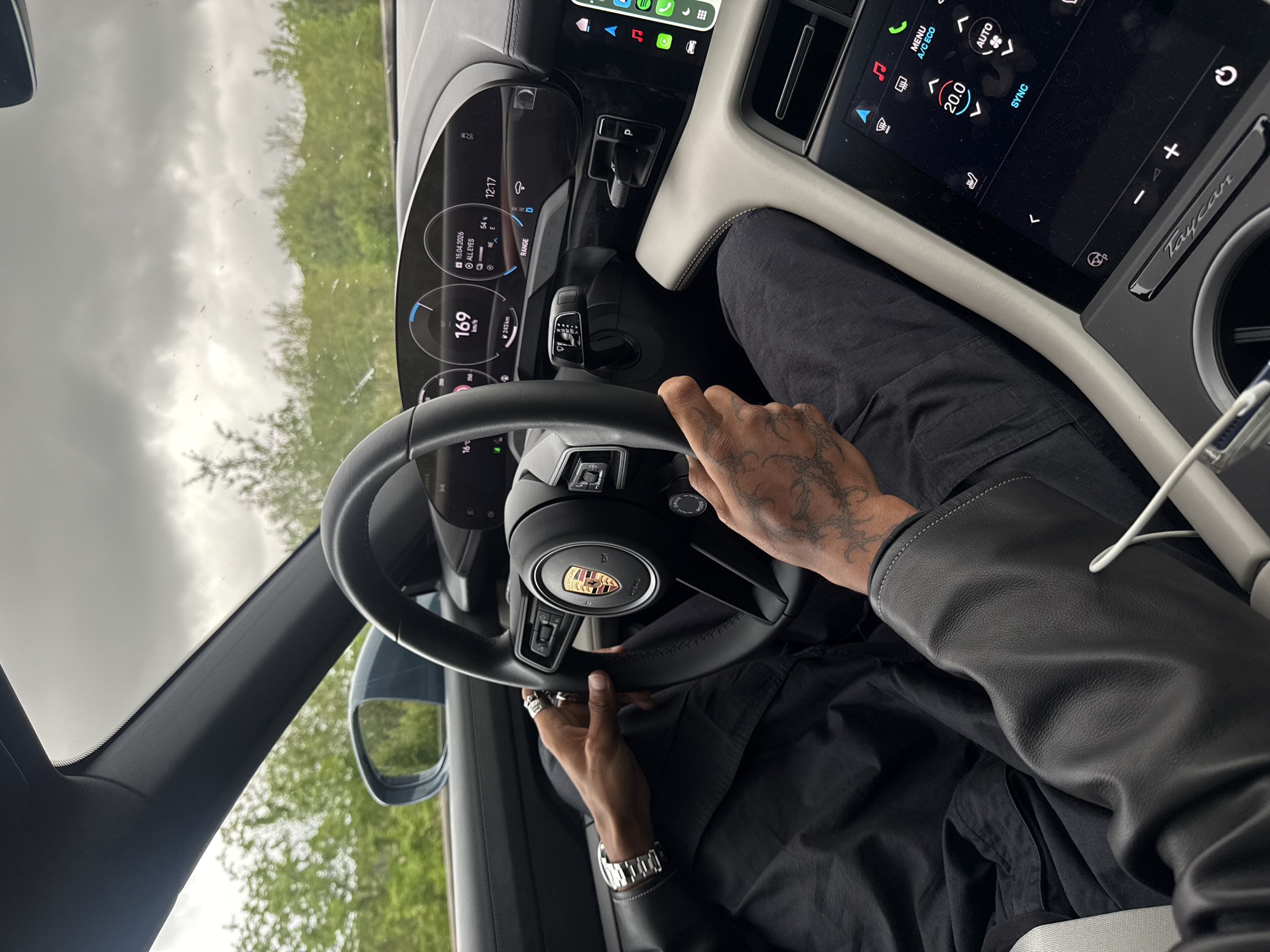 The image shows the interior of a Porsche Taycan, with the driver holding the steering wheel. The driver's hands are visible, adorned with tattoos and rings, and a watch on the left wrist. The car's dashboard is in focus, displaying various information, including a speedometer reading of 169 km/h, the date of April 15, 2026, and a climate control setting of 20.0 degrees Celsius. The car's infotainment screen shows various icons and controls. Outside the car, trees and an overcast sky are visible.