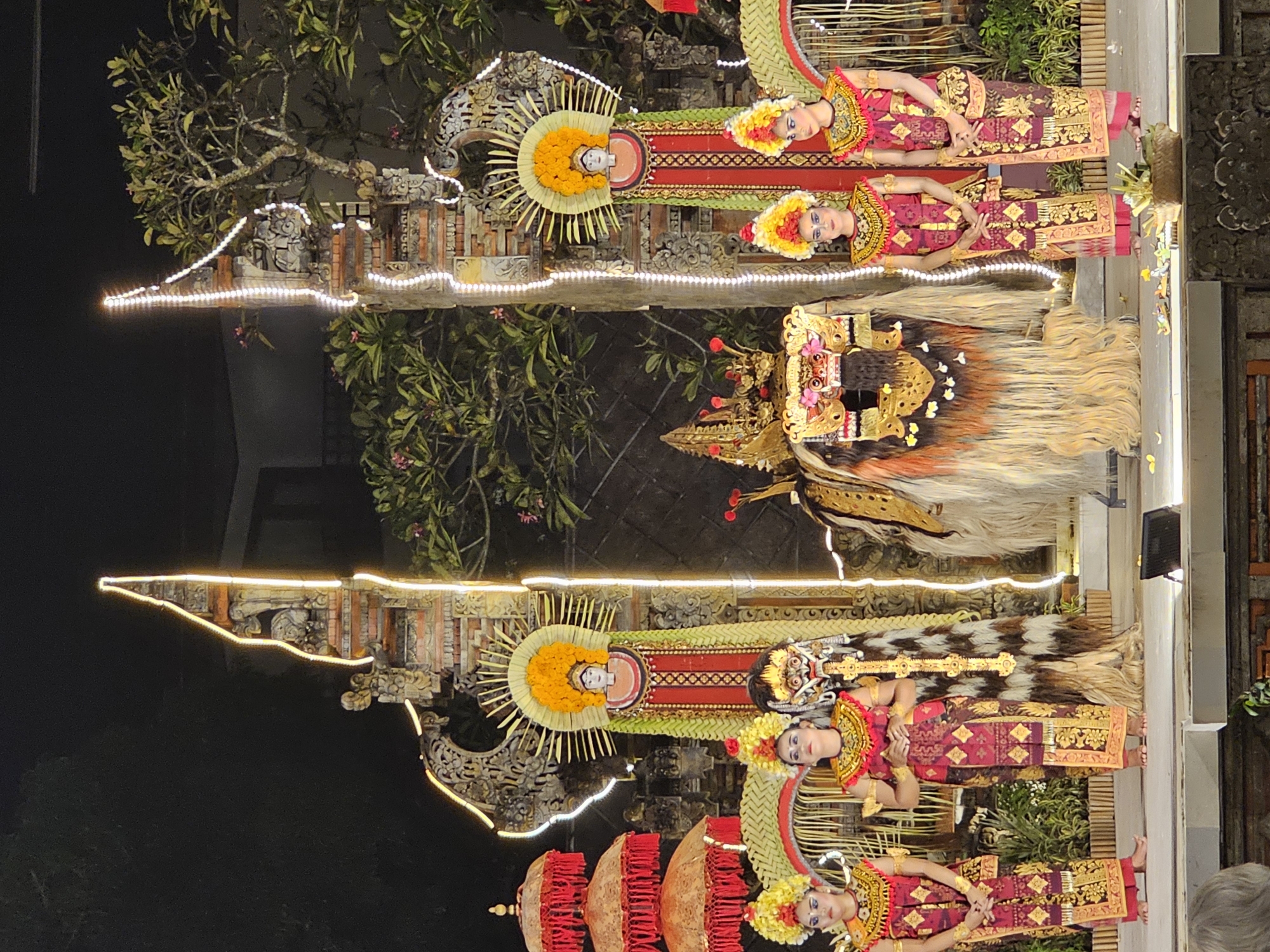 At night in Denpasar, Indonesia, a group of performers are staged outside in front of trees and building exteriors decorated with illuminated white string lights. At least six Balinese dancers are visible in ornate, bright costumes; one dancer is wearing a headdress made of straw or reeds. A Barong costume is in the center of the scene, with a large mask and a long, white and brown mane. The dancers appear to be posing rather than moving dynamically.