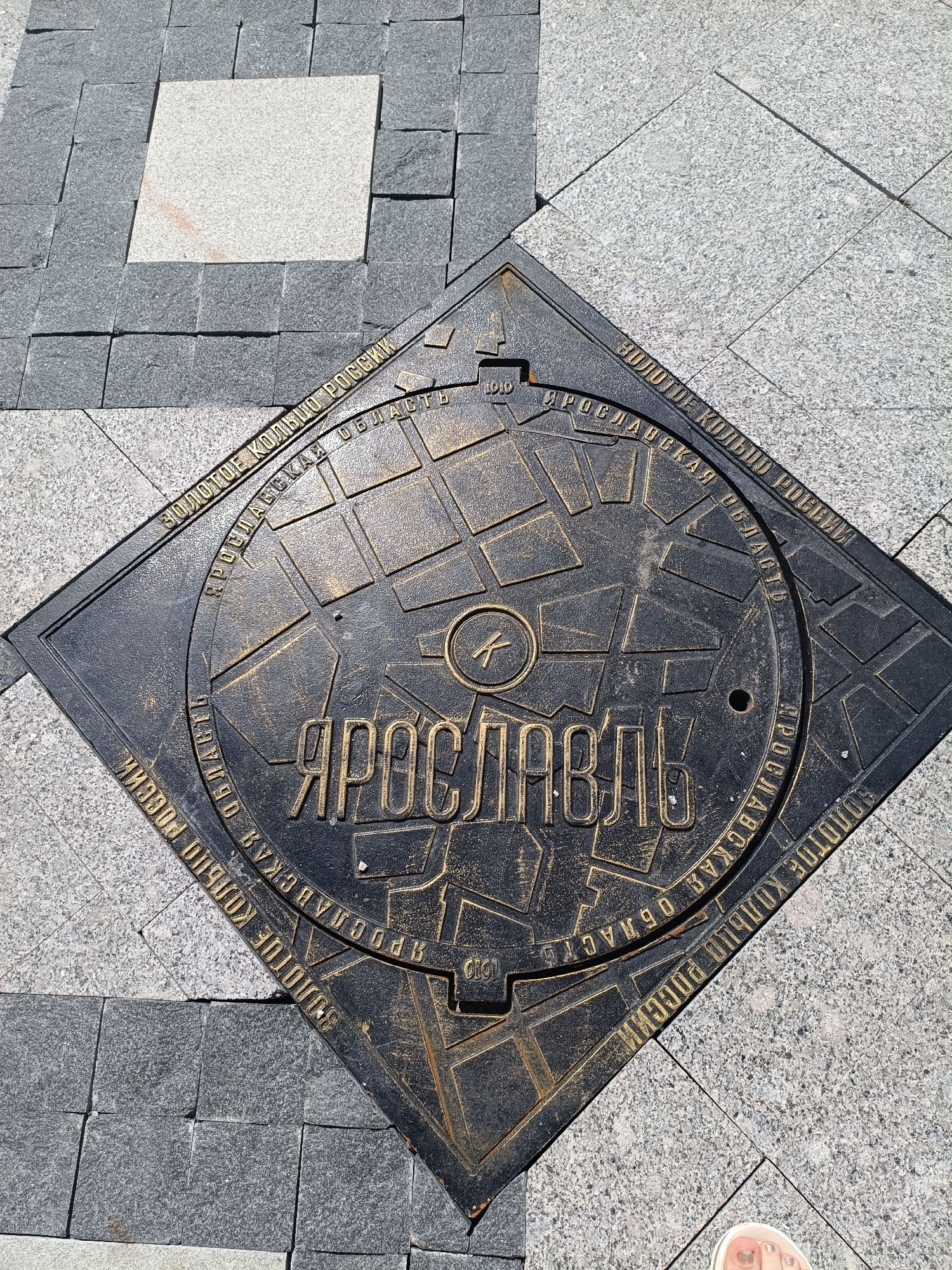 The image shows a square manhole cover on a paved ground, with the cover oriented diagonally. The manhole cover is black with golden lettering and a map-like design. The text on the manhole cover reads "ЗОЛОТОЕ КОЛЬЦО РОССИИ" (Golden Ring of Russia) and "ЯРОСЛАВСКАЯ ОБЛАСТЬ" (Yaroslavl Region). In the center of the manhole cover, the word "ЯРОСЛАВЛЬ" (Yaroslavl) is inscribed in large golden letters. A small "K" in a circle is visible to the left of "ЯРОСЛАВЛЬ". The surrounding pavement is made of grey granite tiles, with a single lighter-colored square tile in the upper left quadrant of the image. A person's foot, with painted toenails, is visible in the lower right corner of the frame, suggesting the photo was taken by someone standing there. The scene appears to be an outdoor public space in the city of Yaroslavl, Russia, likely on a sunny day given the shadows and bright lighting.