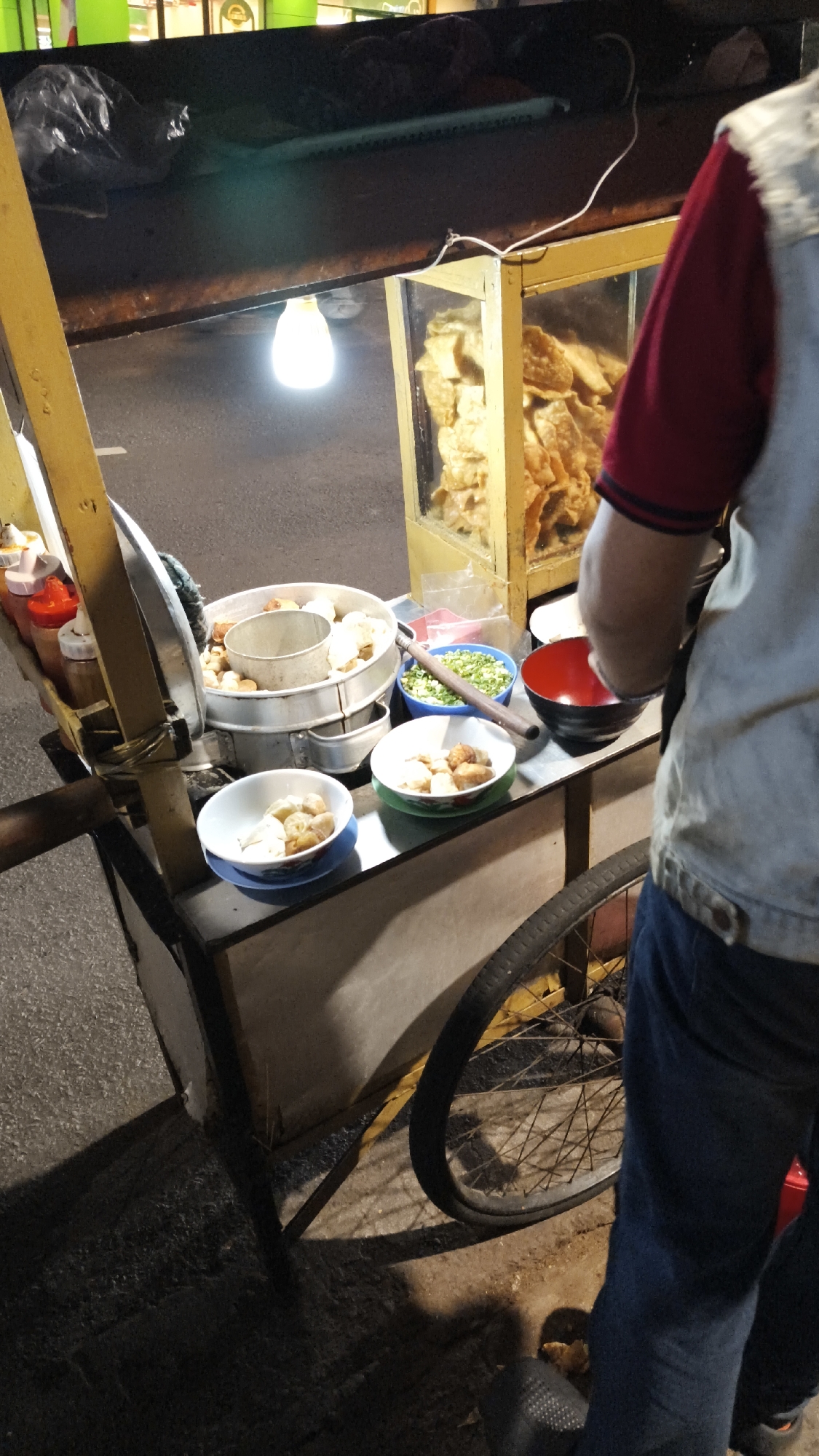 A street food cart is parked on a paved surface at night, likely in South Jakarta, Indonesia. The cart, made of wood and metal, has yellow accents and a wheel at the rear. A single incandescent light bulb illuminates the immediate area. Various food items are displayed on the cart, including what appear to be fried snacks in a glass-fronted display case, and a metal steaming container filled with round food items. There are also bowls containing chopped green onions, fried morsels, and what looks like boiled meatballs. Several condiment bottles are visible on the left side of the cart. A person, partially visible from the shoulder down, is wearing a light-colored vest over a red shirt and blue jeans, and appears to be standing behind the cart, interacting with the food. Shadows are cast on the ground, indicating artificial lighting.