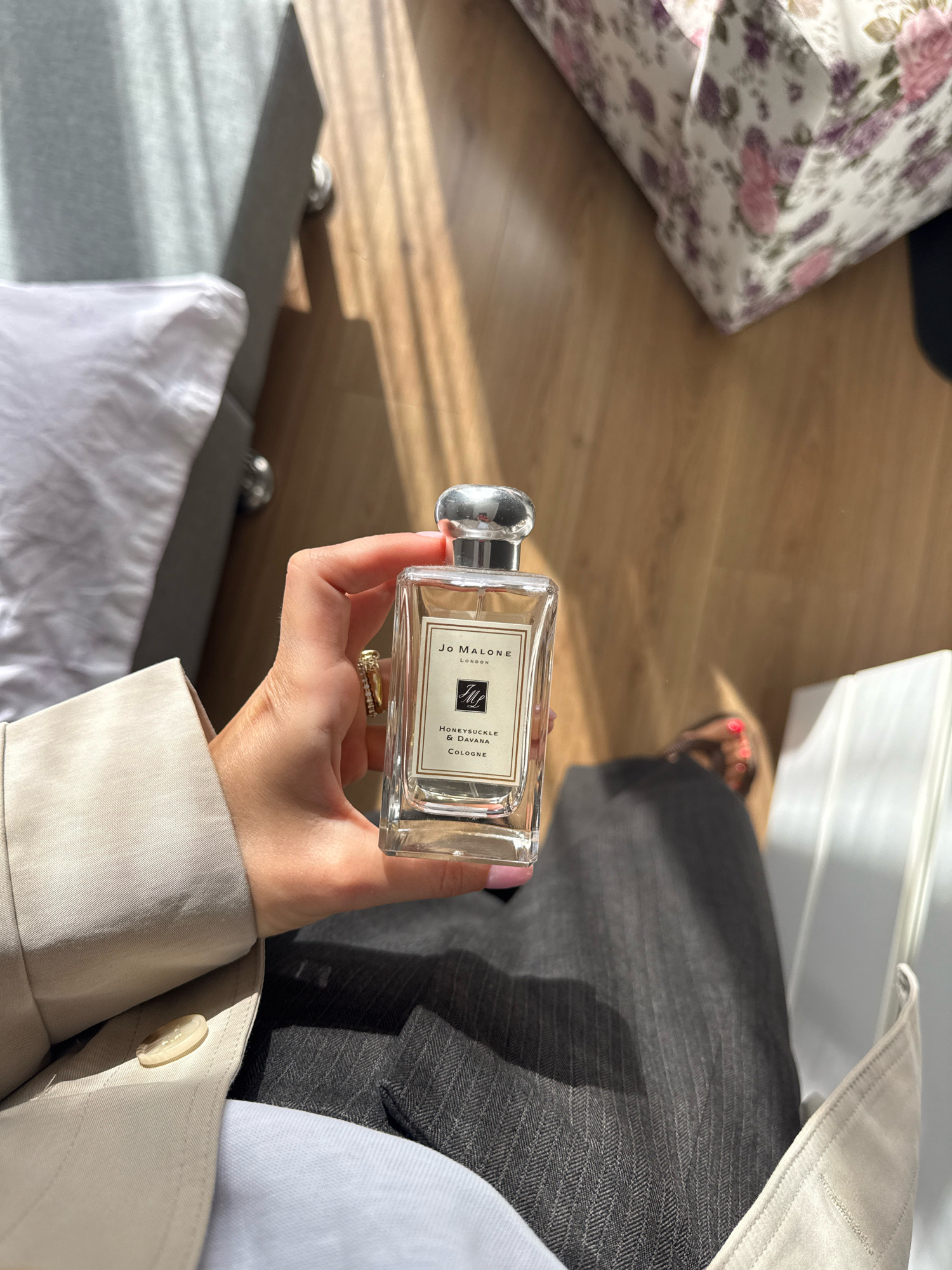 The image shows a close-up of a person's hand holding a Jo Malone London Honeysuckle & Davana Cologne bottle. The person is wearing a light-colored blazer and dark pinstripe pants. The background features a wooden floor with dappled sunlight, a bed with white linens, and a floral-patterned object that appears to be a box or bag. There are no identifiable landmarks or indications of the city of Tbilisi, Georgia in the image.