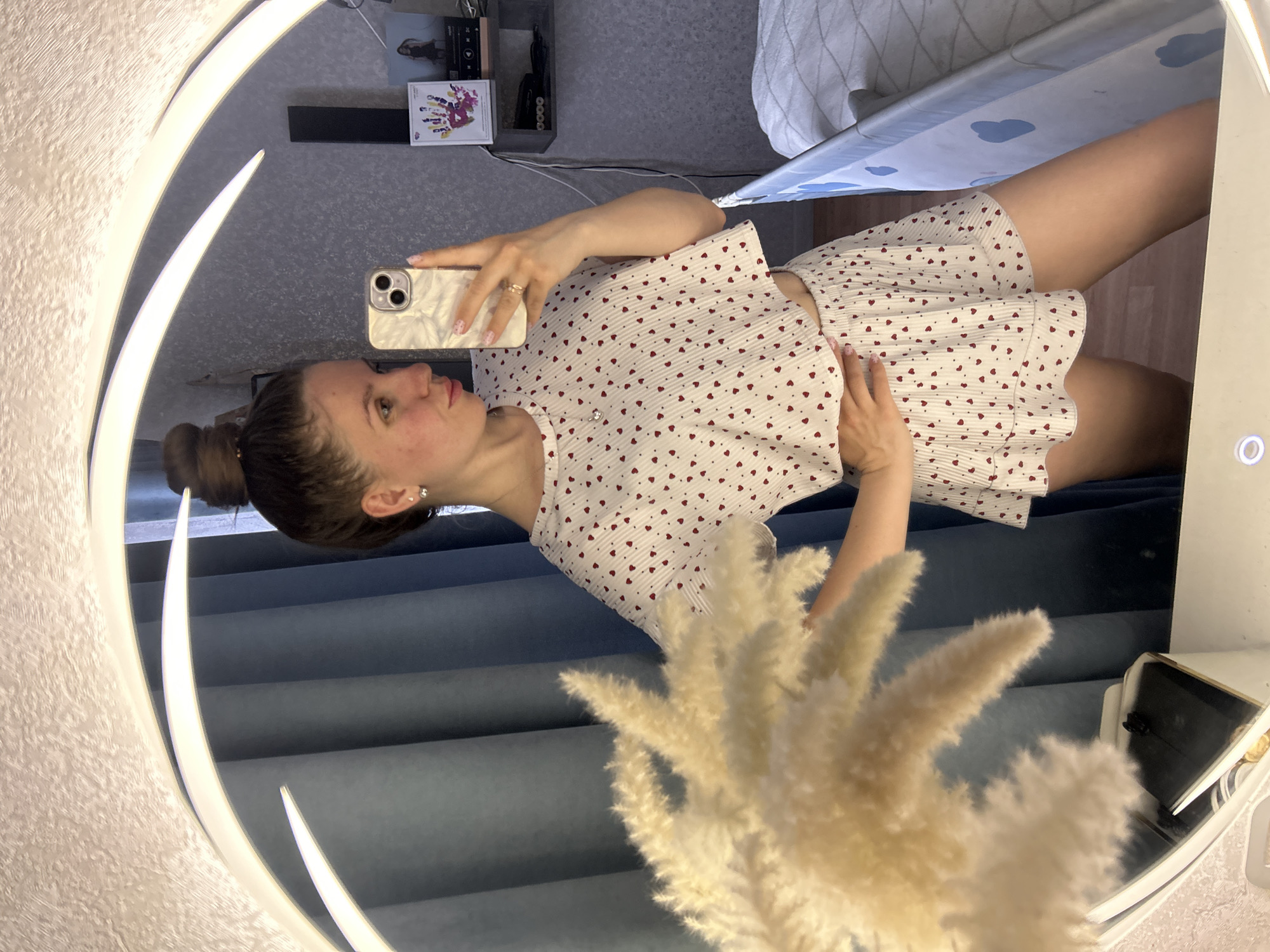 A woman with a bun hairstyle is taking a mirror selfie. She is wearing a white pajama set with red hearts. The mirror is round and illuminated with white light. In the reflection, there is a shelf with various items on it, and part of an ironing board. The background includes a curtain and what appears to be a wall with textured wallpaper. In the foreground, a vase with pampas grass partially obscures the view. There is no indication of the city of Klin, Russia.