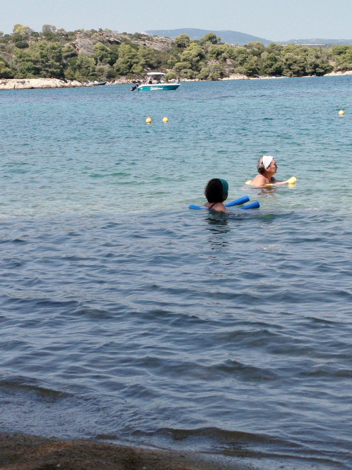 Two individuals are swimming in the ocean in Sithonia Municipal Unit, Greece. The person in the foreground, wearing a green swim cap, is partially submerged and using two blue foam pool noodles. Behind them, an older individual with a white headband is also in the water, with a yellow flotation device. A small boat is visible in the background on the left, and a treed, rocky coastline lines the horizon. The water is clear and calm, with ripples reflecting the light.