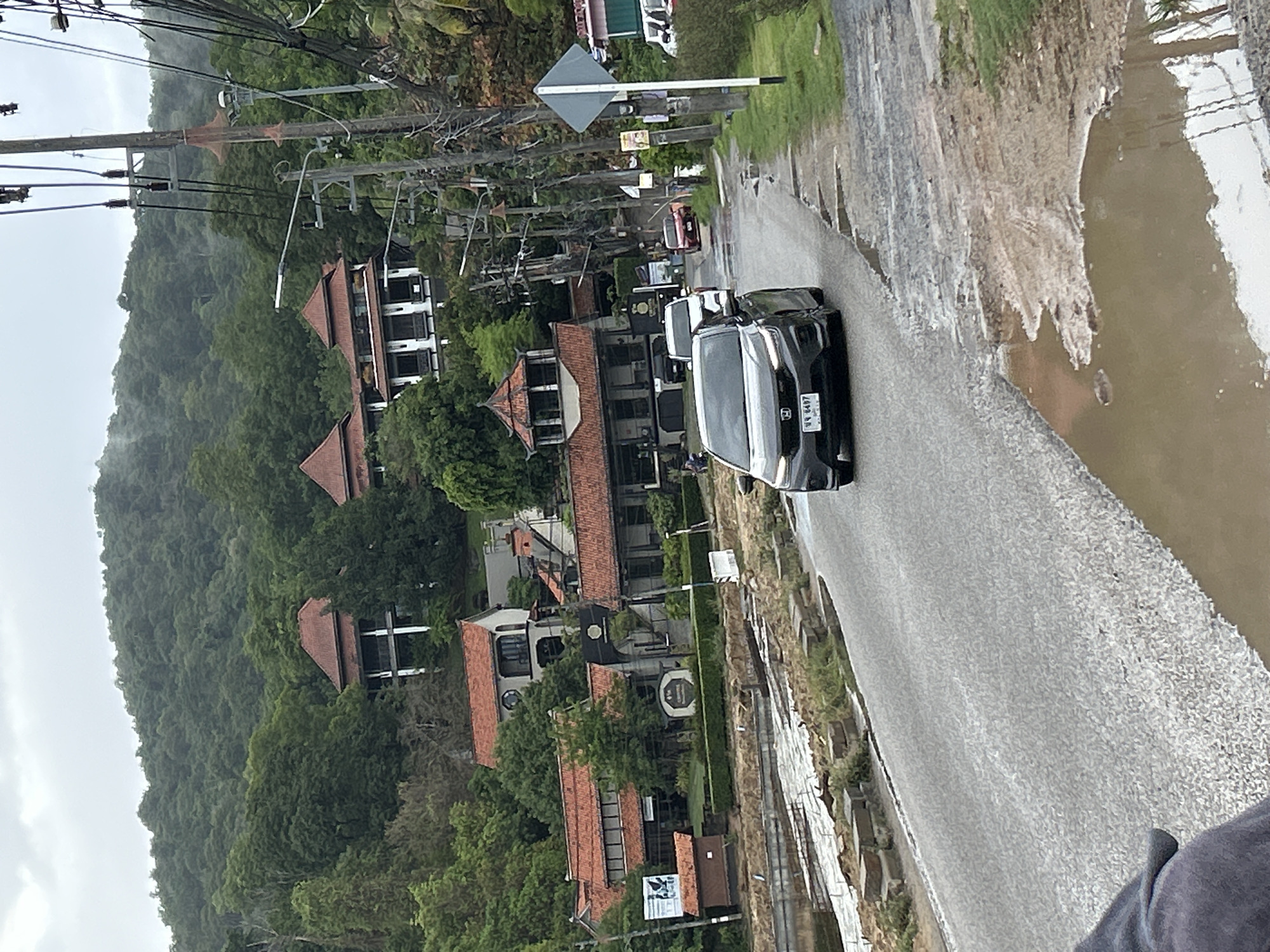 The image depicts a street scene in Rawai, Thailand, during or after a rainstorm. The road is wet and has standing water in the shoulder. On the right side of the road, a black car is traveling in the direction of the viewer. Several houses and buildings with red-tiled roofs are visible on the left side, nestled among lush green trees. In the background, a densely forested hill rises up. Power lines stretch across the top of the image.