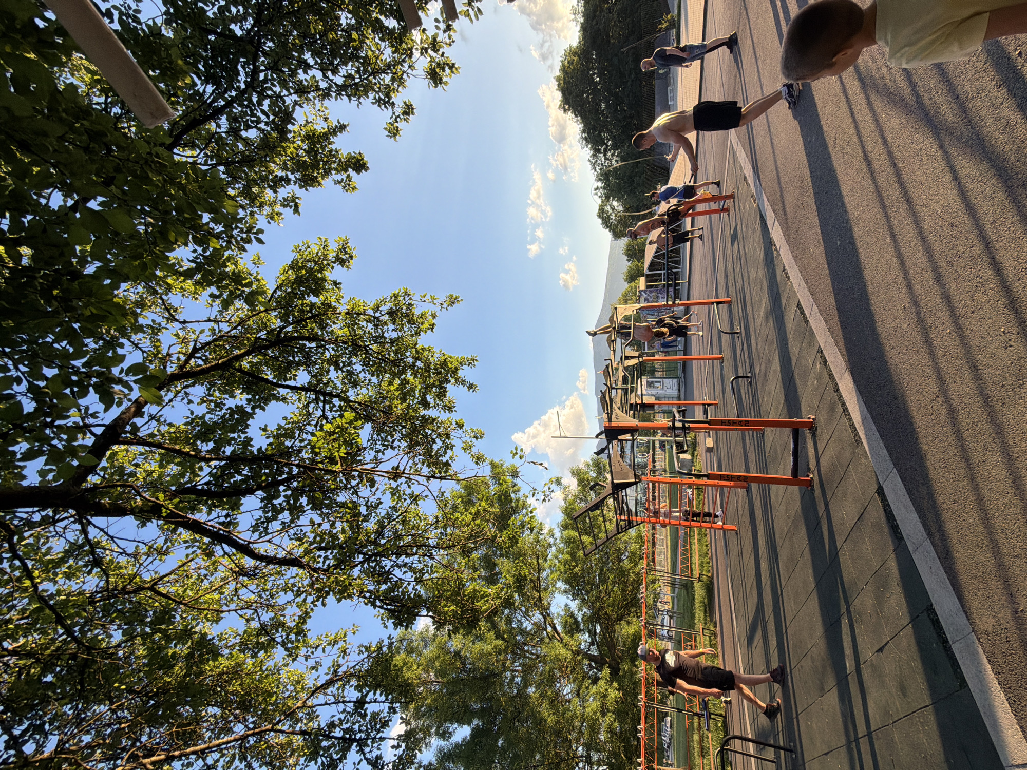 The image shows a park in Sofia, Bulgaria, with exercise equipment and people using it. The image is taken from a low angle, looking up at the equipment and the people exercising. There are trees on either side of the frame, and the sky is visible in the center. The exercise equipment includes bars, ladders, and other structures. The people are engaged in various exercises, such as pull-ups, dips, and stretching. The environment is sunny and outdoors.