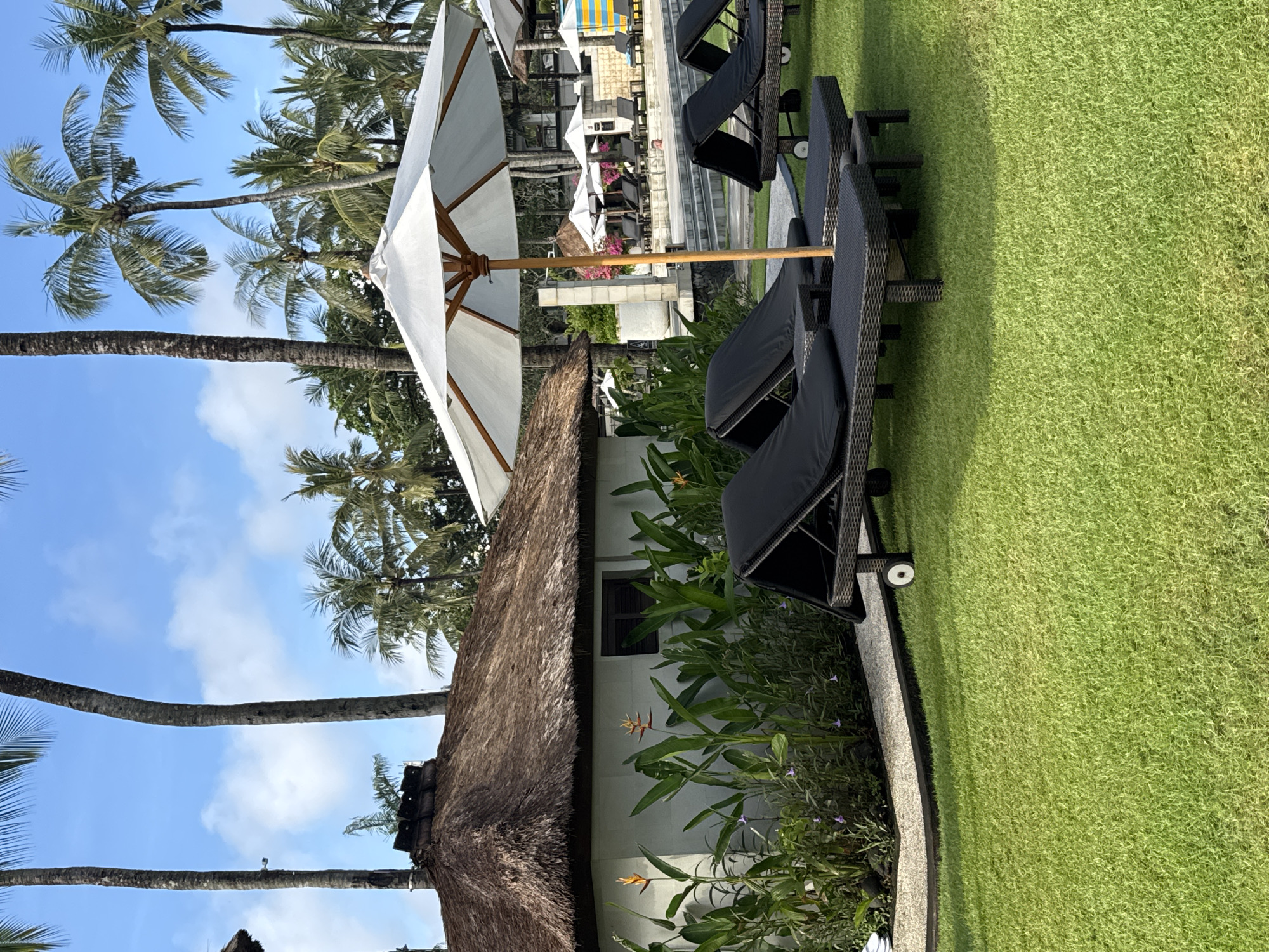 This vertical image depicts a tranquil tropical outdoor setting, likely a resort or hotel in Kuta Selatan, Indonesia. The scene is captured in bright daylight under a clear blue sky adorned with scattered white clouds.
The right side of the frame features a well-maintained, vibrant green lawn. Along the edge of this lawn, several black woven lounge chairs, some with dark cushions and wheels, are neatly arranged, suggesting an area for relaxation. A narrow, light grey concrete border separates the lawn from a lush garden bed. This bed is filled with various green tropical plants, including broad-leafed foliage and flowering species, notably some distinctive orange and yellow bird of paradise flowers, as well as smaller purple blossoms.
On the left, a small building with white walls and a rustic thatched roof, typical of tropical architecture, is visible. Numerous tall palm trees, characterized by their slender trunks and green fronds, dominate the upper and left portions of the image, reinforcing the tropical environment. A large, white sun umbrella with a wooden central pole stands prominently, ready to provide shade.
In the background, hints of a larger leisure area are visible, including what appears to be a swimming pool area with additional sun umbrellas, more lounge seating, and light-colored architectural elements like steps or platforms. No people are present, emphasizing the serene and inviting atmosphere of the unoccupied leisure space. The image conveys a sense of peace and an ideal vacation destination.
