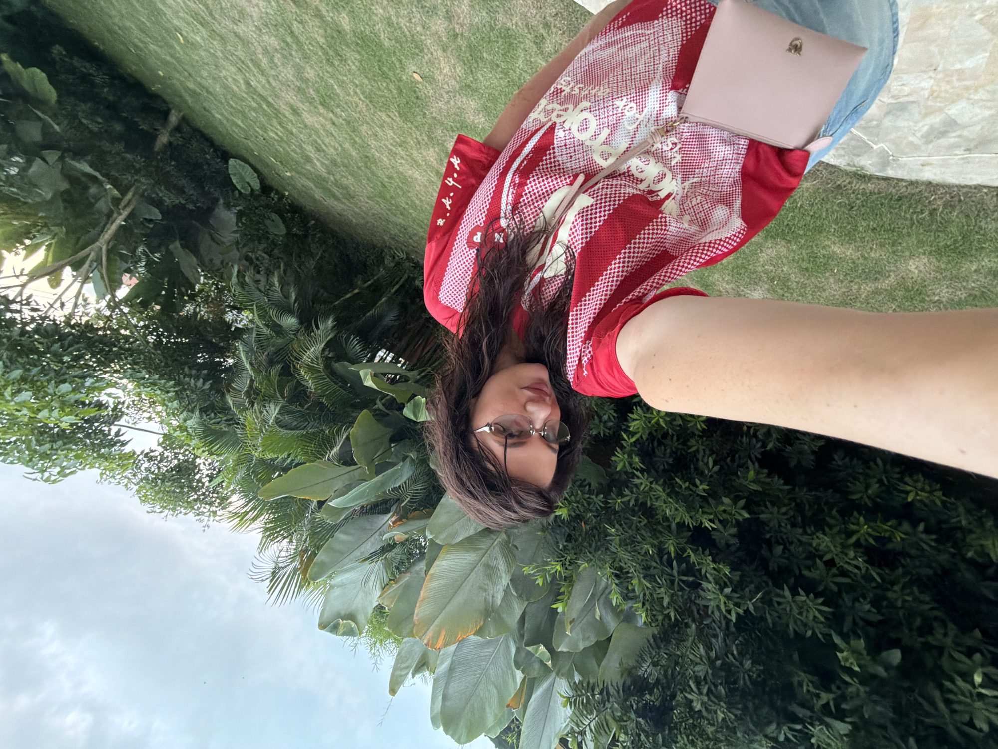 A selfie-style image, taken outdoors during the daytime under a cloudy sky. The primary subject is a person, likely female, with long dark brown hair, wearing eyeglasses. They are looking directly at the camera with a neutral expression.
The person is wearing a red and white patterned shirt, which features white stylized text that appears to be "SOLO" or similar words repeated as a design. On the left sleeve, there are vertical red characters on a white background that resemble Japanese or Chinese script. They are also wearing blue jeans. A light pink or beige clutch purse with a small gold emblem is resting on their lap.
The setting is lush and green, indicating a tropical or subtropical environment, consistent with North Kuta, Indonesia. The background is dominated by various large-leafed tropical plants, ferns, and trees. There is a patch of well-maintained green grass to the right of the person, and a light-colored stone or concrete path/ledge is visible under the grassy area. The overall scene suggests a relaxed outdoor environment, possibly a garden, resort, or park.
