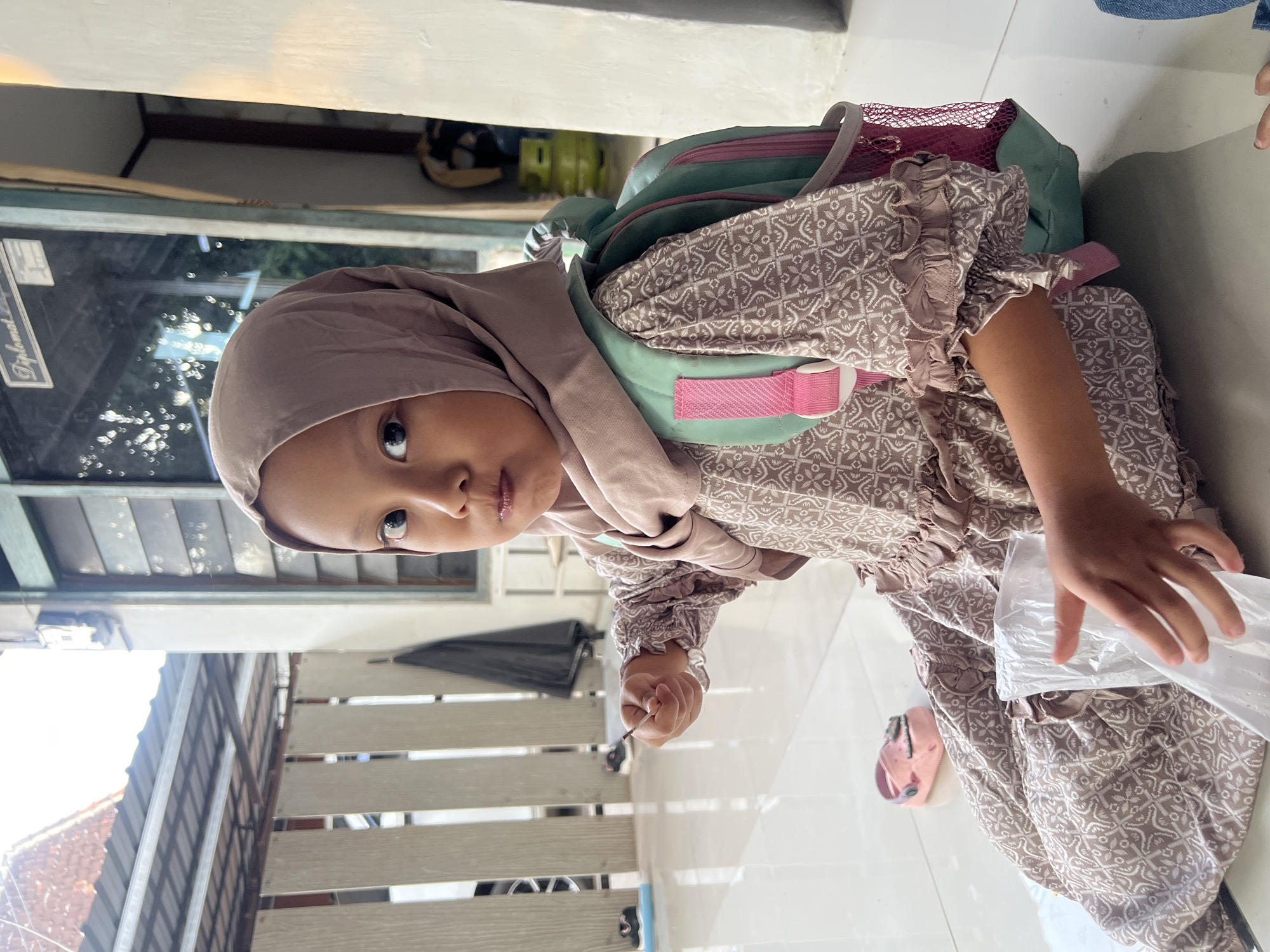 A young girl wearing a light brown hijab and a patterned dress sits on a tiled floor outdoors. She has a light turquoise and pink backpack on her back and is holding a white plastic bag in her hands. To her left, on the floor, is a pink sandal. In the background, a metal awning covers an outdoor area, and a window with wooden frames is visible. A green gas cylinder is also present in the background, and a black umbrella leans against a fence. The scene appears to be in a residential area, potentially in Kecamatan Pati, Indonesia, given the architectural style and the gas cylinder which is common in Indonesian households. The lighting suggests it is daytime, with sunlight casting shadows.