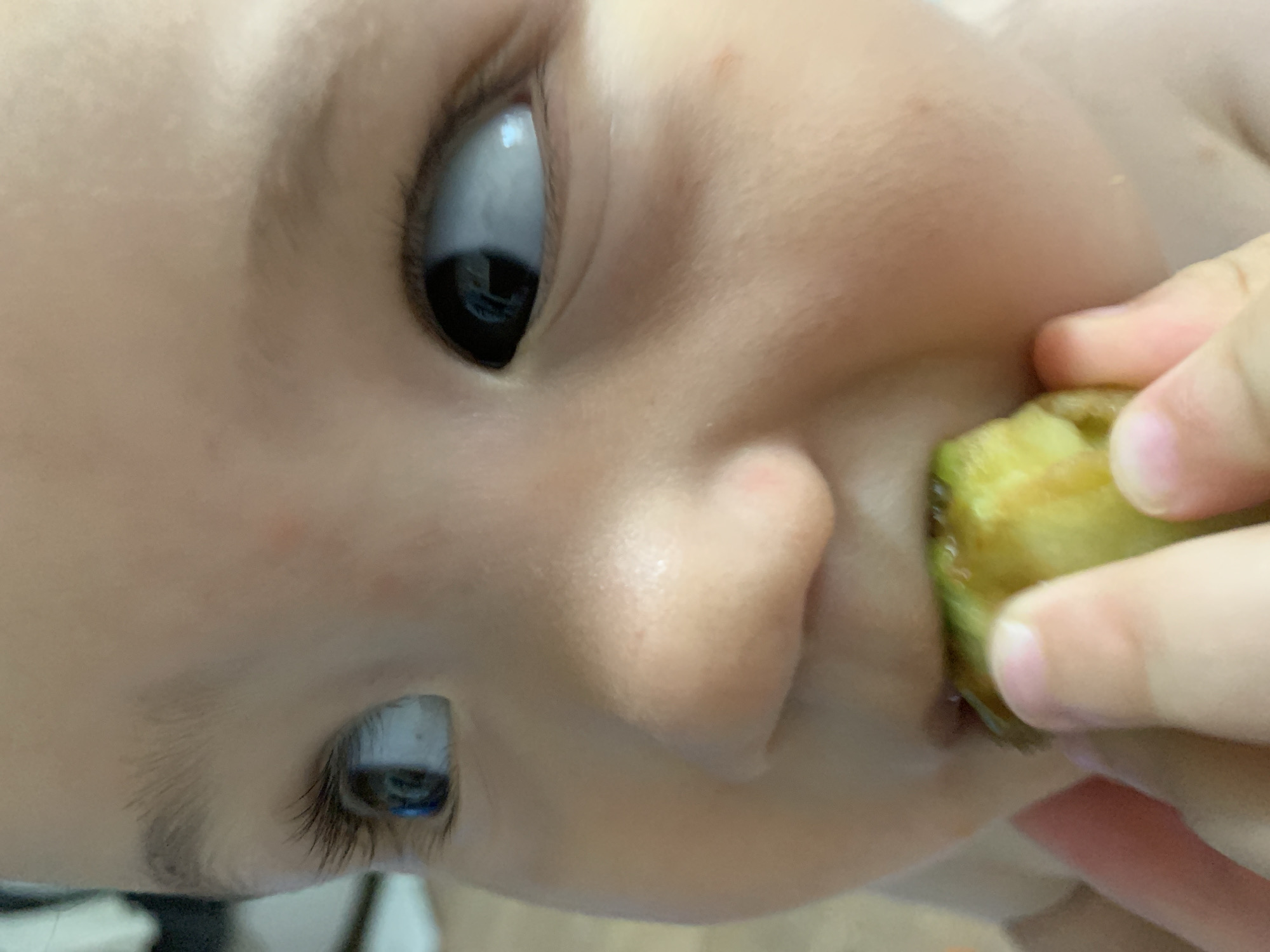 A close-up, slightly blurry, eye-level shot shows a child's face, with the focus on the child's eyes and mouth. The child's left eye is looking to the left and slightly upward, and the right eye is looking to the right. The child's mouth is partially open, and they are holding a piece of green food, possibly a vegetable, in their mouth. The child's skin is light-skinned, and they have dark eyelashes and eyebrows. The background is out of focus and appears to be an indoor setting with neutral colors. There are no identifiable landmarks or features that would indicate a specific location in Chaikovsky, Russia.