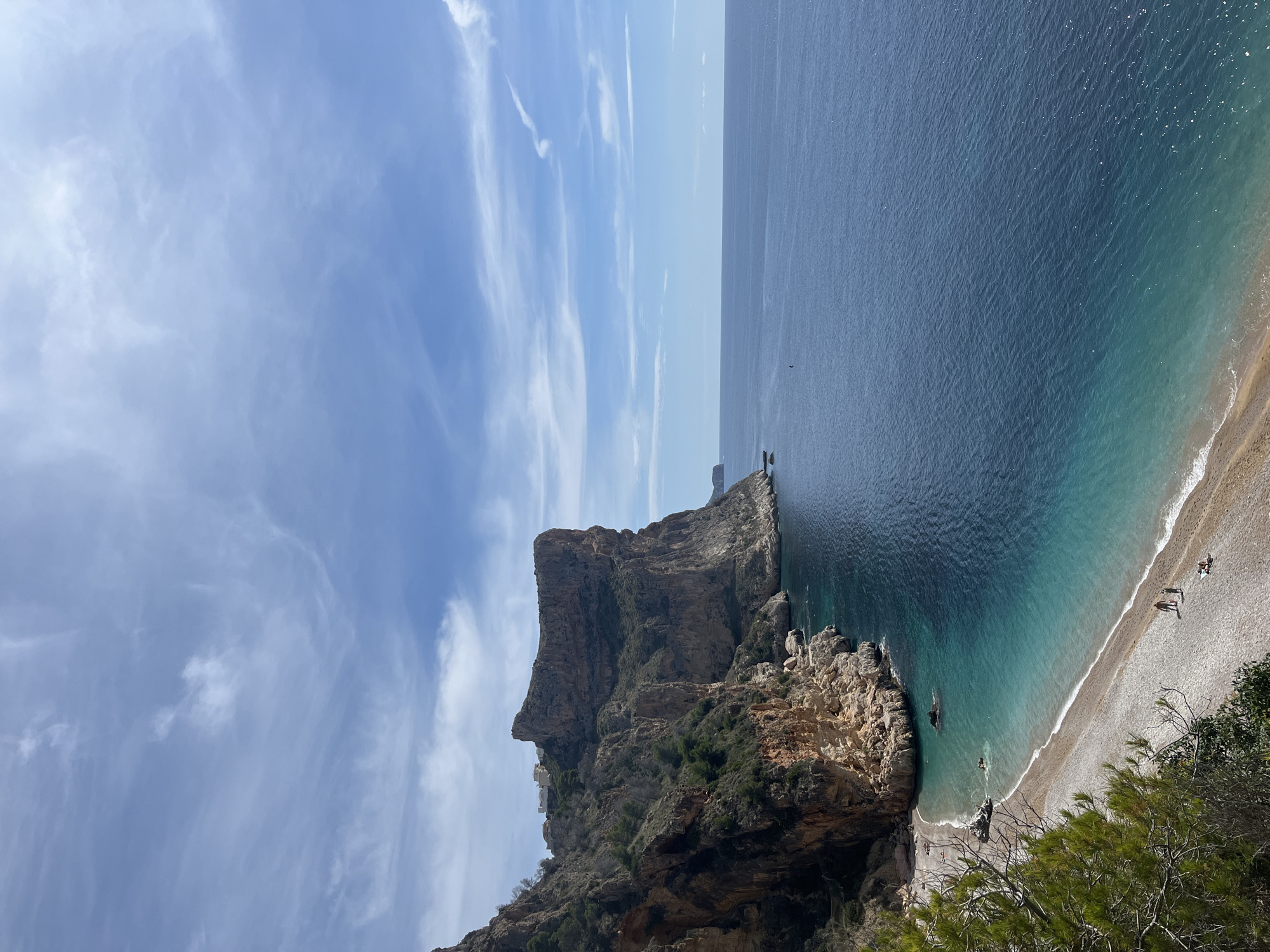The image captures a coastal scene in El Poble Nou de Benitatxell/Benitachell, Spain. A large, steep cliff dominates the left side of the image, dropping sharply into the turquoise water. The cliff face has varying rock colors and textures, with sparse vegetation clinging to its slopes. The ocean transitions from a lighter turquoise near the shore to a deep blue further out. A beach of small, light-colored pebbles lines the coast. Several people are visible on the beach. The sky is mostly clear, with scattered wispy clouds.