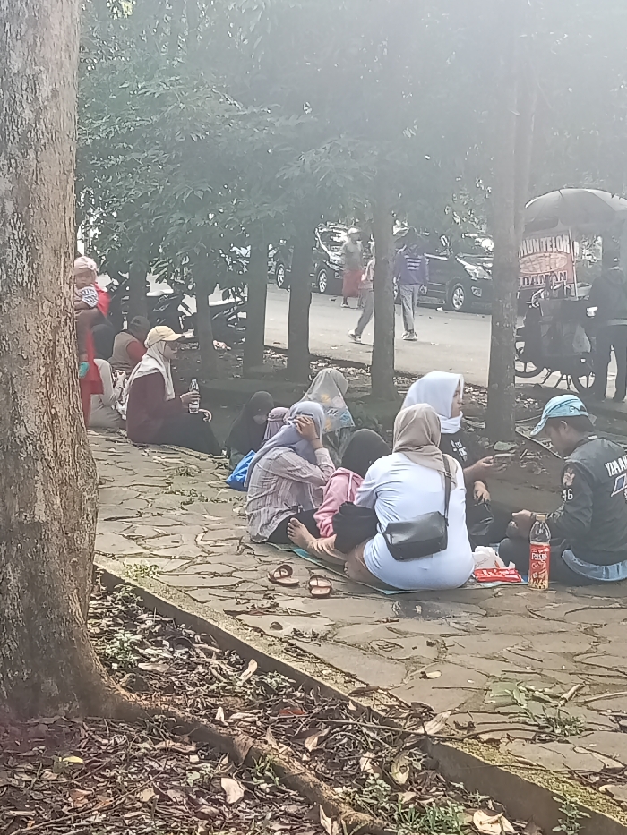The image depicts an outdoor scene in Singaparna, Indonesia, where a group of people are gathered, predominantly women, many wearing headscarves (hijabs). They are seated on a stone-paved path under the shade of trees, suggesting a casual rest stop or social gathering.
In the foreground, a large tree trunk is visible on the left, with its roots extending into the earthy ground. On the paved area, several individuals are present. On the far left, a woman in a red top is holding a child, partially obscured by the tree. Beside her, another woman wearing a light-colored hijab and a cap holds a clear bottle. Further to the right, a cluster of people are seated on what appears to be mats or blankets. One woman in a white top wears a black cross-body bag, while another in a striped shirt has her hands to her head. A man in a blue cap and a dark jacket, bearing the visible text "YAMAHA," sits facing right, with a red-labeled bottle (likely "FRESTEA") and other small items in front of him. Several pairs of sandals are scattered on the path nearby.
In the mid-ground and background, more trees line what appears to be a road where cars are parked. People are seen walking or standing. On the far right, a food cart mounted on a motorcycle is partially visible, with text like "OMUN TELOR" and "DANAN" faintly discernible on its canopy, possibly advertising street food.
The scene takes place during the daytime, characterized by bright, somewhat hazy sunlight filtering through the trees. The overall atmosphere appears relaxed, with individuals engaged in resting, eating, or conversing.
