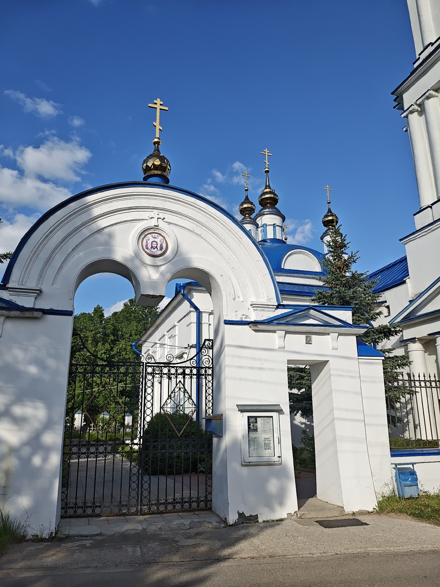 The image shows an Orthodox church in Мамонтово, Russia. The church is white with blue accents on the roof and trim. It features multiple golden domes topped with Orthodox crosses. An arched entranceway with a gate in the foreground leads to the church grounds. A religious icon is visible within the arch. A notice board is attached to one of the gate pillars. Green trees are seen through the gate. The sky is blue with scattered clouds.