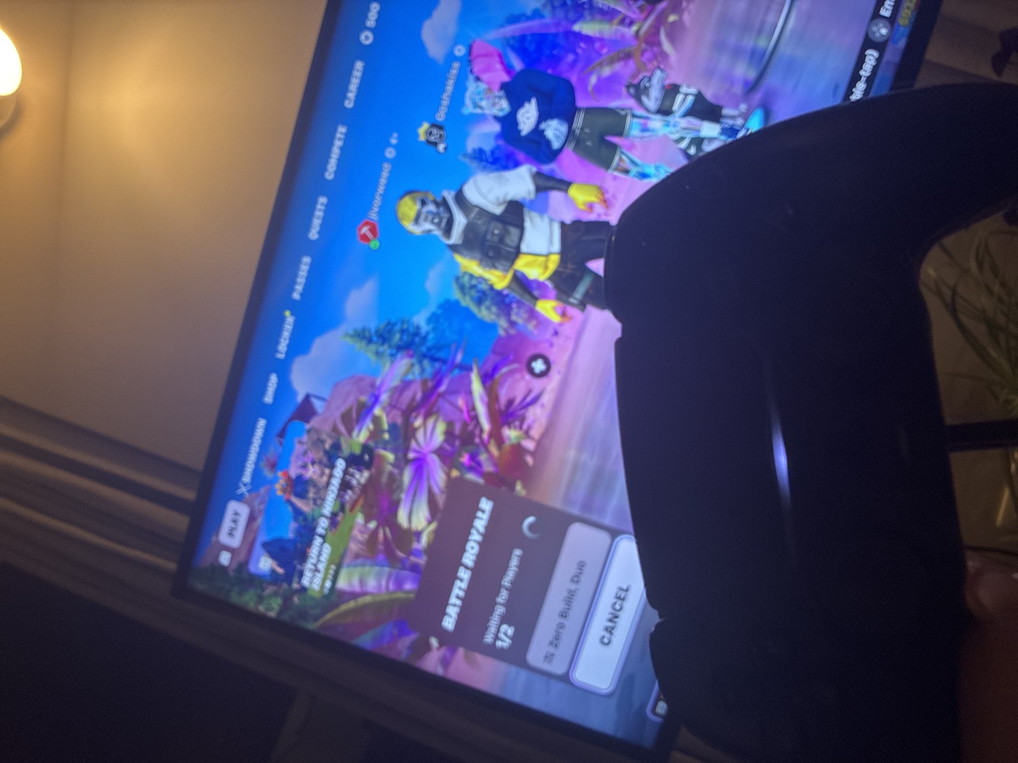 An angled, slightly blurry shot captures a television screen displaying the menu of a video game, presumably Fortnite, with a dark game controller obscuring part of the right side. The screen shows two player avatars; one in a yellow and black outfit with a yellow helmet, and another in a blue hoodie and dark pants. The background of the game is a vibrant, colorful landscape featuring purple and green foliage.
The game's interface is visible, with top menu options including "PLAY", "XSHOWDOWN", "SHOP", "LOCKER", "PASSES", "QUESTS", "COMPETE", "CAREER", and "500". The primary focus on the screen is the "BATTLE ROYALE" mode, which indicates "Waiting for Players" and a status of "1/2" for a "Zero Build, Duo" match. A "CANCEL" button is also present. The player names "jiverweed" and "Goshakiss" are visible above the avatars. Other text includes "RETURN TO NINJAGO ISLAND".
In the upper left corner, a light fixture casts a warm glow on an off-white wall, suggesting an indoor setting and possibly evening time. The image quality is somewhat soft, particularly the details of the controller and the light.