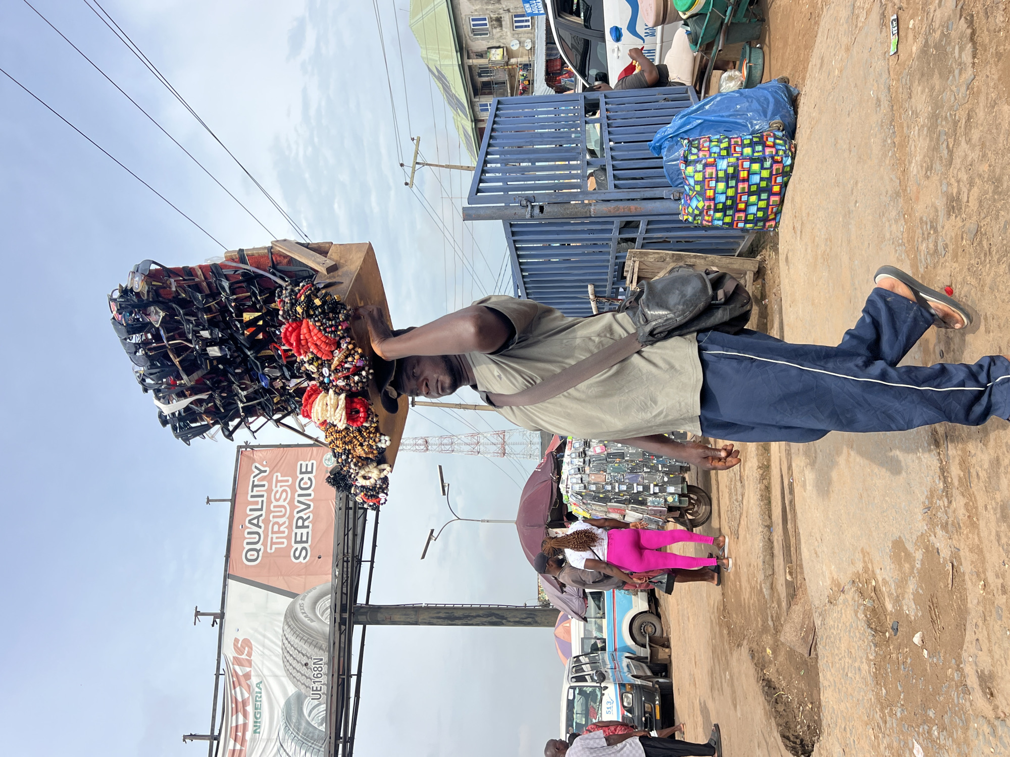 A Black man walks carrying a wooden crate filled with sunglasses and beaded accessories on his head. He is wearing a green t-shirt and blue pants, with a black satchel slung across his chest. The man is walking on a dirt road with potholes, in what appears to be a market. In the background, there are several vehicles, including a blue and white bus, and stalls with umbrellas. A large billboard advertising "Maxxis Nigeria" and "Quality, Trust, Service" is visible in the distance, along with power lines and utility poles. The sky is overcast. The scene is set in Onitsha, Nigeria.