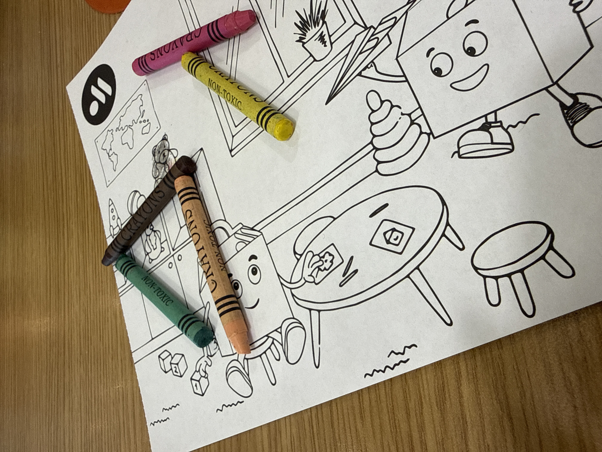 A close-up shot depicts a child's coloring page with several crayons placed on top. The image features a partially completed coloring sheet with cartoon-style illustrations of furniture, a box-like character with a smiling face, and a stack of toys. Four crayons—pink, yellow, brown, green, and orange—are scattered over the paper. The words "NON-TOXIC" are visible on some of the crayons. The coloring page seems to be resting on a wooden surface, likely a table. There is no information about the location in the city of Dmitrov, Russia, in the image.