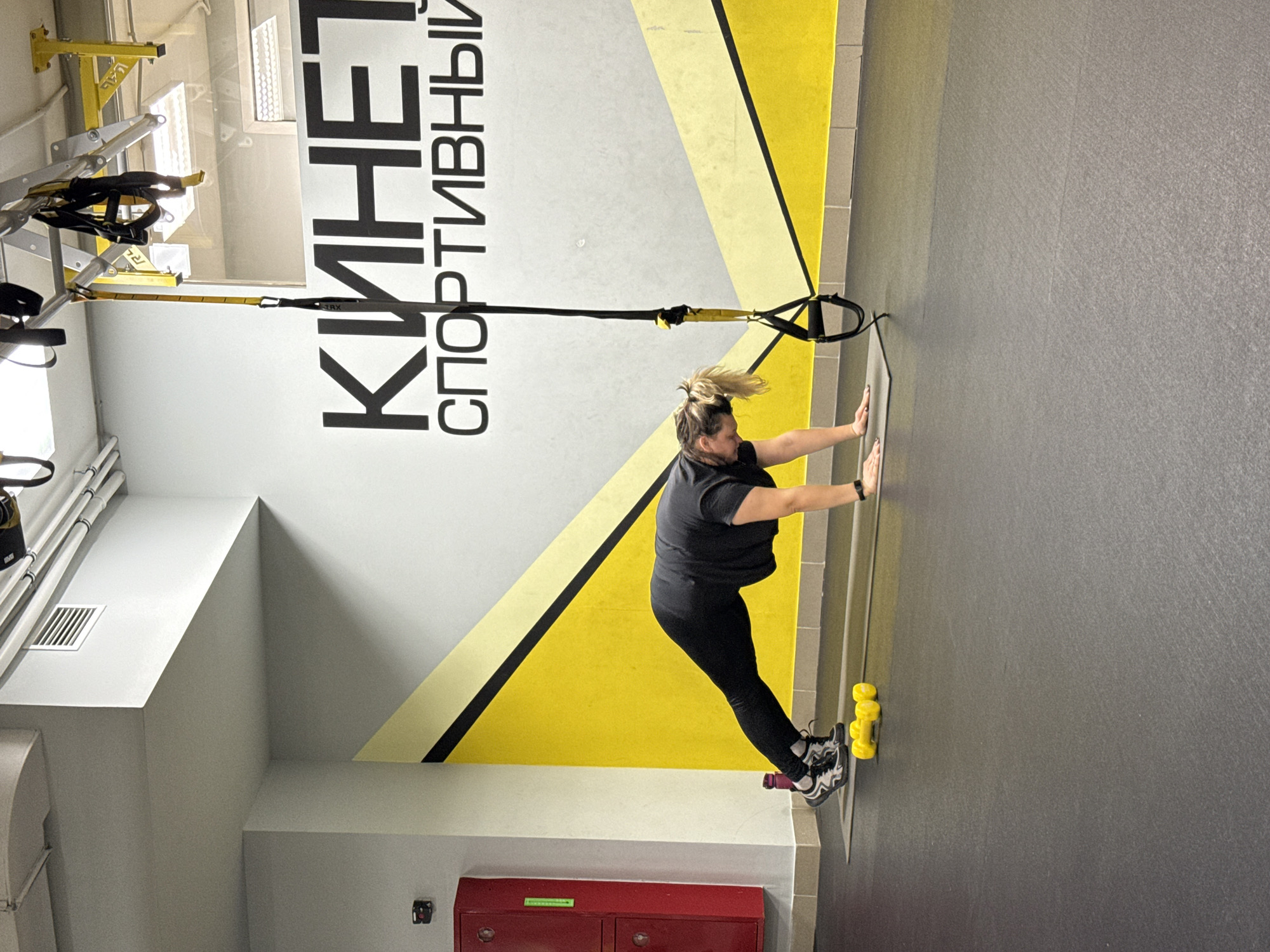 The image depicts a woman performing a wall push-up exercise using TRX suspension straps in what appears to be a gym. She is wearing a black top and leggings, with her feet braced against the wall. The wall has a yellow and black triangular design. The location is identified by the sign "KINET SPORTEVNBI" on the wall, indicating the city of Chaikovsky, Russia. In the background, there is gym equipment and part of a red cabinet. The gym has a gray floor and white walls.