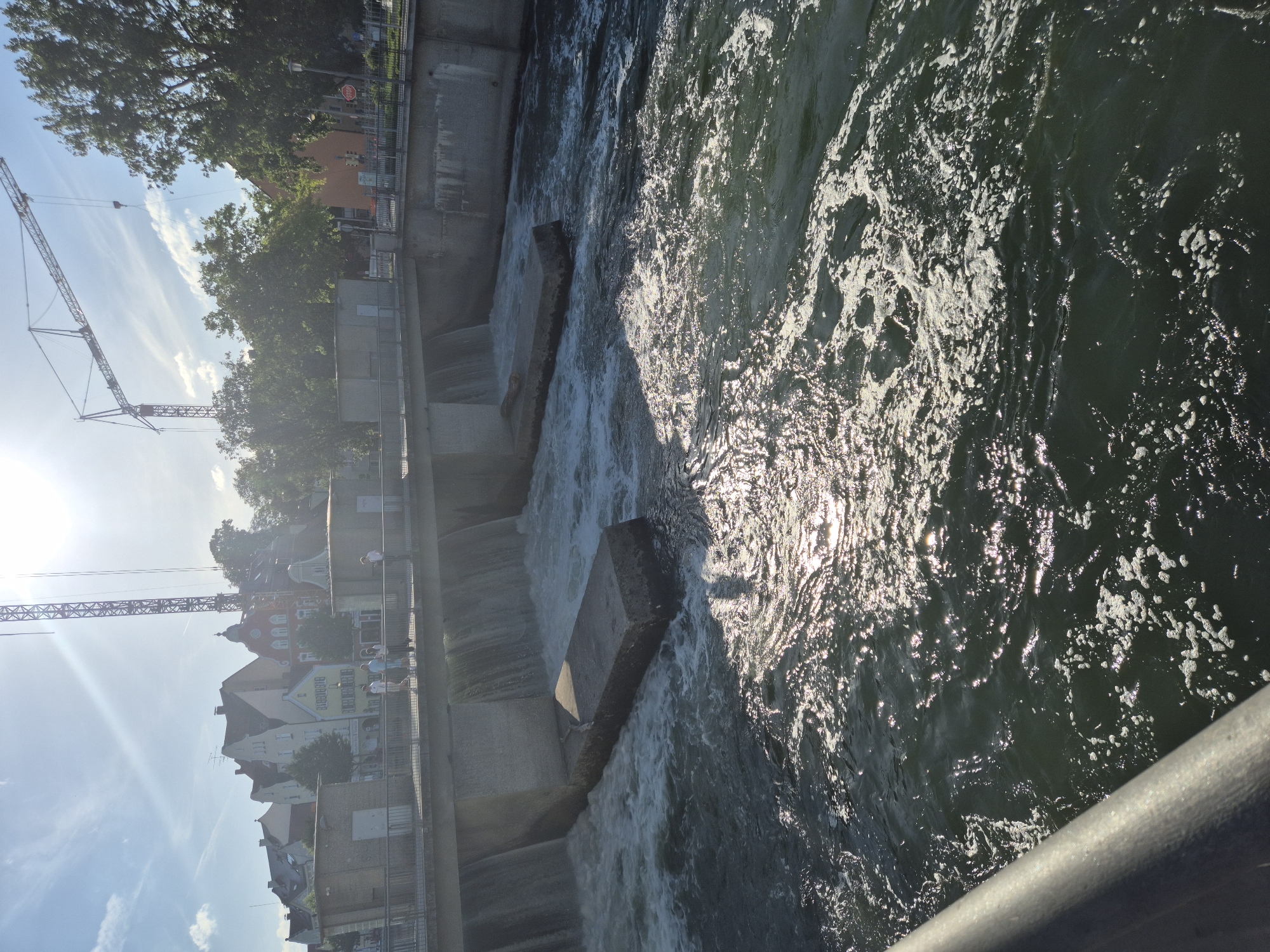 The image depicts a scene at the Isar river in Landshut, Germany, specifically at the dam and weir structure. The sun is bright, casting a glare on the water and creating lens flare in the upper right quadrant.
In the foreground, the dark, turbulent water of the Isar flows rapidly, with white foam and spray generated by the cascading water. A concrete barrier or railing runs along the bottom left edge of the frame.
The central part of the image shows the weir, a man-made structure designed to control water flow. Water is actively spilling over the edge of the weir, creating multiple streams of falling water. In front of the weir, large, rectangular concrete blocks protrude from the water, serving as obstacles or part of the weir's construction.
Beyond the weir, a bridge or walkway is visible. On this walkway, several people are standing and walking, appearing to observe the river or the surrounding area. They are dressed in casual, light-colored clothing.
In the background, a townscape with buildings of varying architectural styles is visible. These buildings have pitched roofs and appear to be typical of European urban architecture. Behind the town, large green trees are present, and further in the distance, construction cranes are prominent against the sky, suggesting ongoing development or infrastructure work in the city. The sky is blue with scattered white clouds.