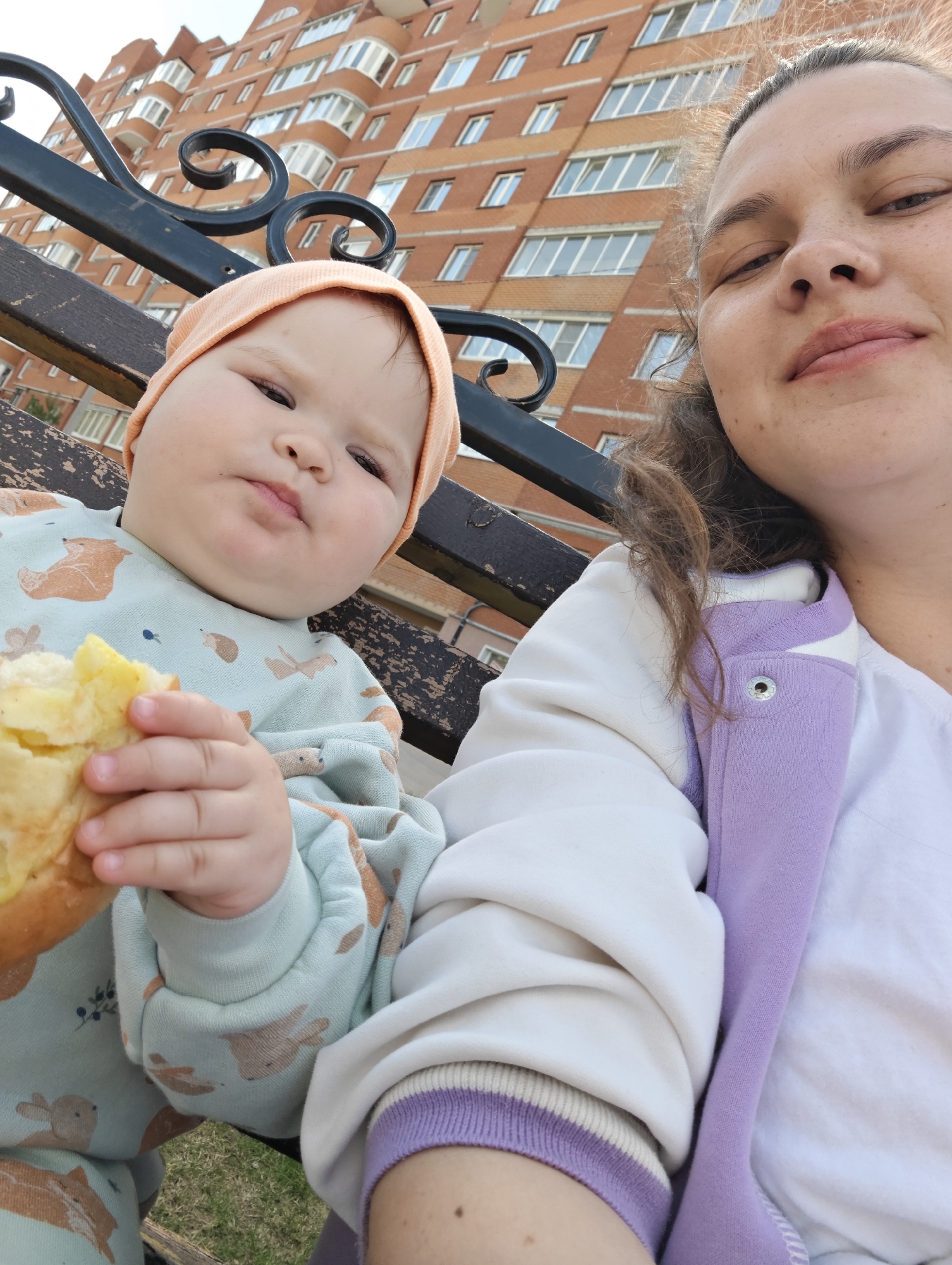 A woman and a baby are sitting on a bench outdoors in Chaikovsky, Russia. The woman is on the right side of the frame, close to the camera, and the baby is on the left. The baby is wearing a light blue patterned outfit and a peach-colored hat, and is holding a piece of yellow food in their left hand. The woman is wearing a white shirt and a white jacket with a purple trim. The background shows a multi-story brick building with many windows, suggesting an urban setting. A black wrought iron bench is visible between the people and the building.