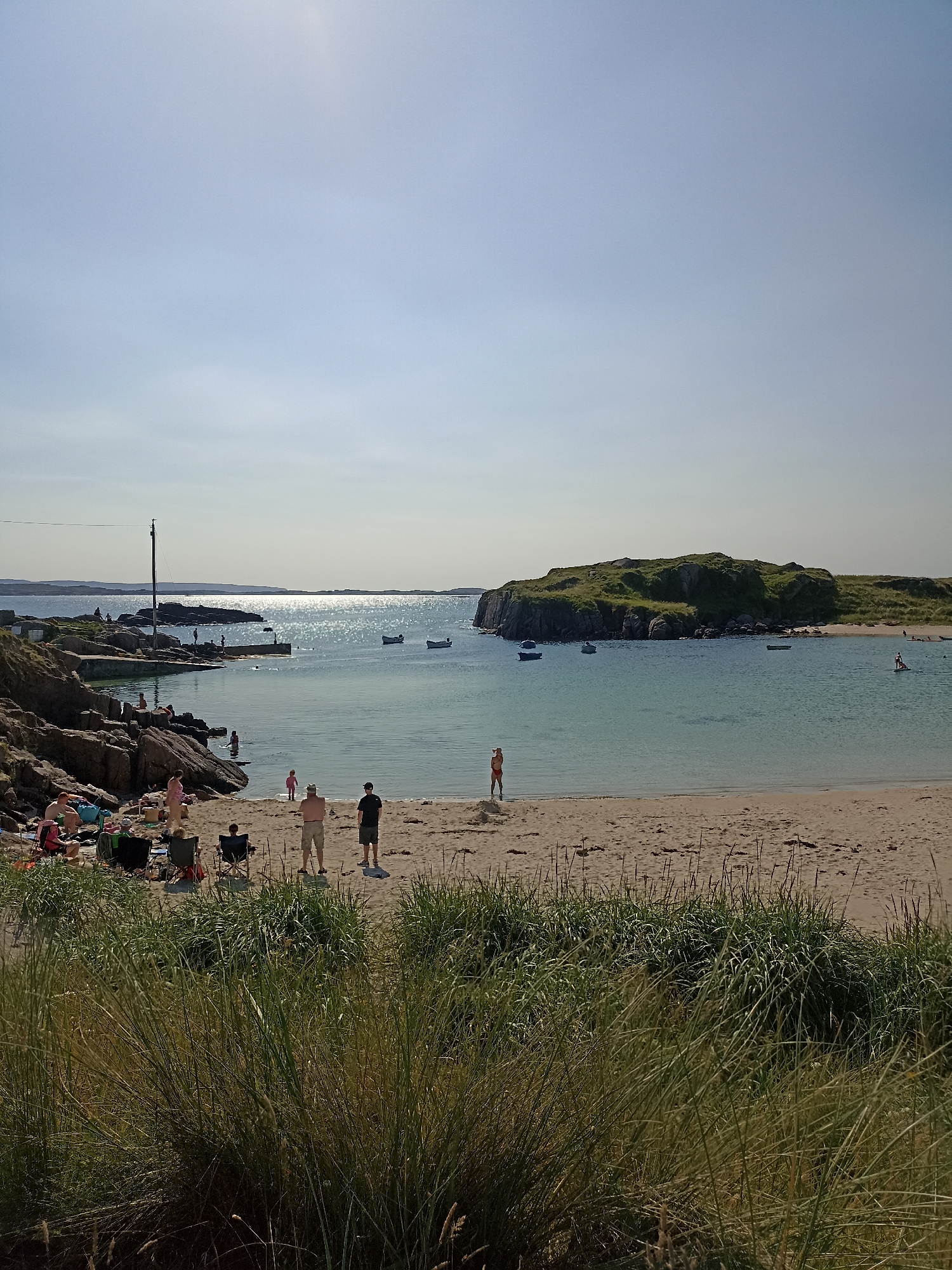 This is a vertical, eye-level, full-shot photograph of a beach scene in Carnboy, Ireland. The sky is a pale, clear blue with a slight haze in the distance. The sun glints off the calm, light blue ocean. In the foreground, a sandy beach is partially obscured by tall, green grass. Several people are scattered across the beach. On the left side, near some rocky outcrops and a small stone pier, a few people are sitting in beach chairs and towels, and others are wading in the shallows. Further into the water, several small boats are moored. In the mid-ground, a small, grassy island with rocky edges rises from the water. On the beach in the mid-ground, two men are standing, and a child is walking towards the water. A woman in an orange swimsuit is standing alone on the sand in the mid-ground. In the background, the coastline of Ireland is visible as a faint, distant line of hills under the hazy sky. The overall scene depicts a relaxed, sunny day at the beach.
