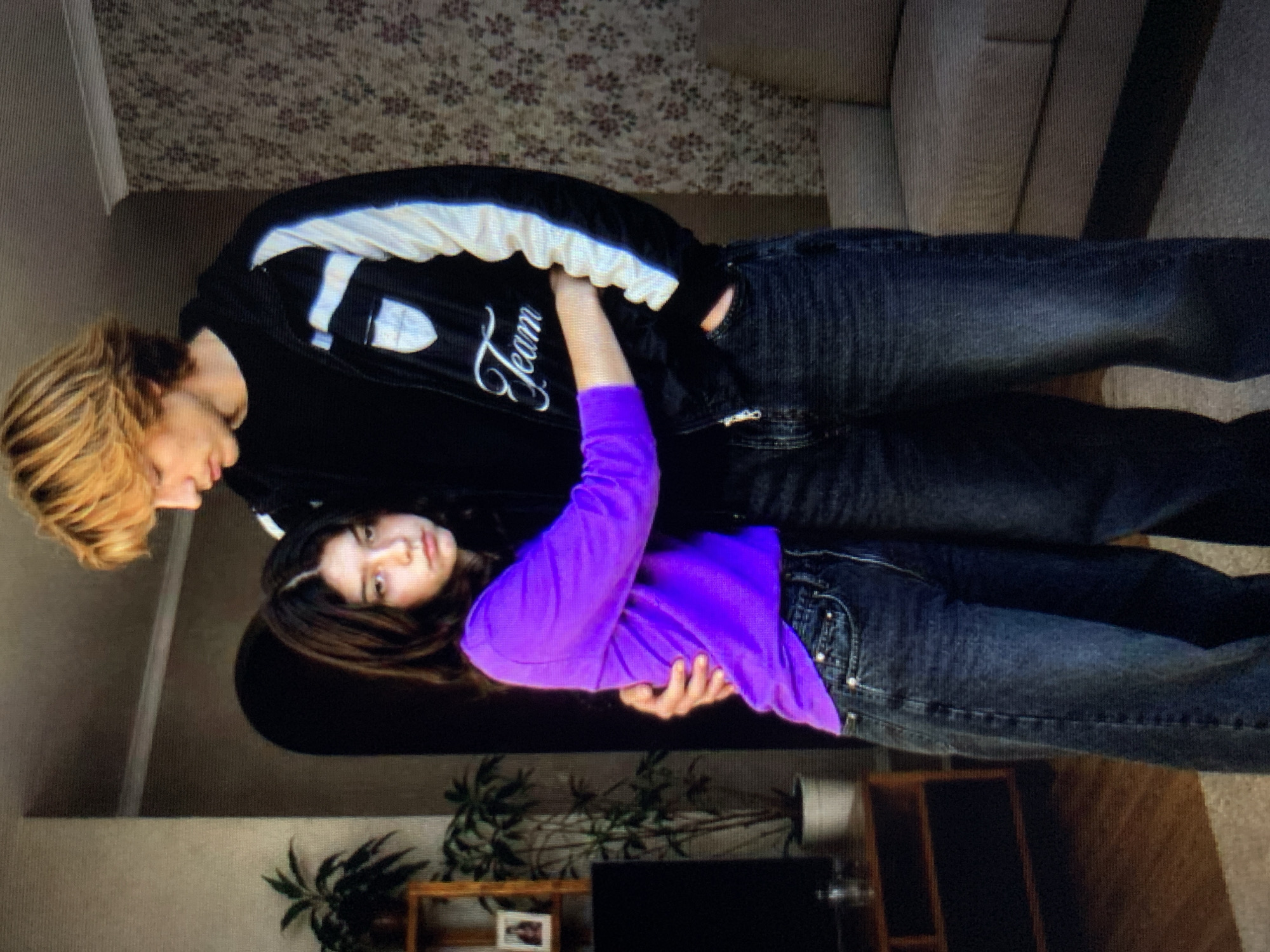 A young woman with dark, wavy hair and a purple long-sleeved shirt is embracing a young man with blonde, shaggy hair. The man is wearing a black jacket with white stripes and the word "Team" embroidered on it, over a black shirt, and dark jeans. The woman is wearing dark wash jeans. They are standing in what appears to be a living room. In the background, there is a floral patterned wallpaper, a couch, a doorway with an arched opening, and a wooden shelf with plants and a framed picture. A portion of a television and a wooden entertainment stand are visible to the left. The floor is carpeted. The scene suggests an indoor, domestic setting, possibly in Lymanka, Ukraine, though no specific city or country is identifiable from the visual information provided.