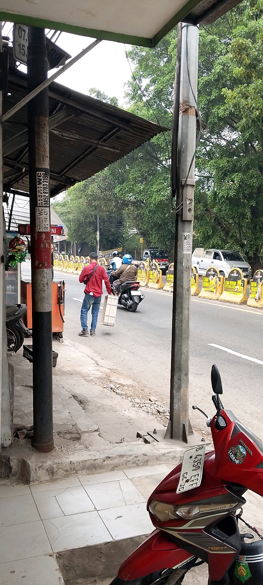 The image depicts a street scene in Depok, Indonesia. In the foreground, a red and black Honda scooter is parked. To the left, a person in a red long-sleeved shirt and blue jeans walks away from the camera, carrying a white plastic crate. They are on a sidewalk, which appears to be cracked and under a covered structure with a metal roof supported by pillars. Behind the person, a motorbike with a rider wearing a blue helmet is moving on the road. The road is lined with yellow temporary traffic barriers. Beyond the road, there are trees with lush green foliage. The sky is overcast. Various signs and stickers are visible on the pillars, including one with numbers "07.15." and another with text and a phone number. A silver pickup truck is visible on the road in the background, behind the barriers.