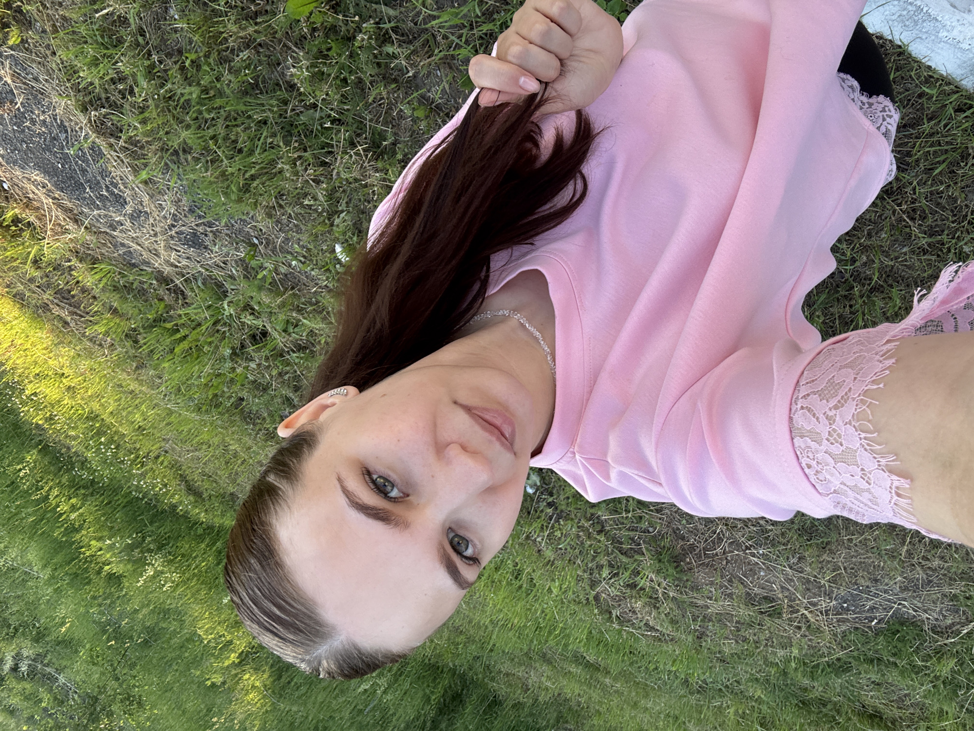 A woman with long dark hair is lying on the grass, looking up at the camera. She is wearing a pink long-sleeved shirt with lace detailing on the sleeves. Her hair is pulled back in a ponytail. The grass is green and appears to be slightly overgrown in some areas. A path or road is visible to the left of the frame, with dry grass and debris along its edge. The sun is shining, casting shadows on the grass, suggesting it is daytime. The woman's expression is relaxed and pleasant. The image appears to be a selfie, taken outdoors.