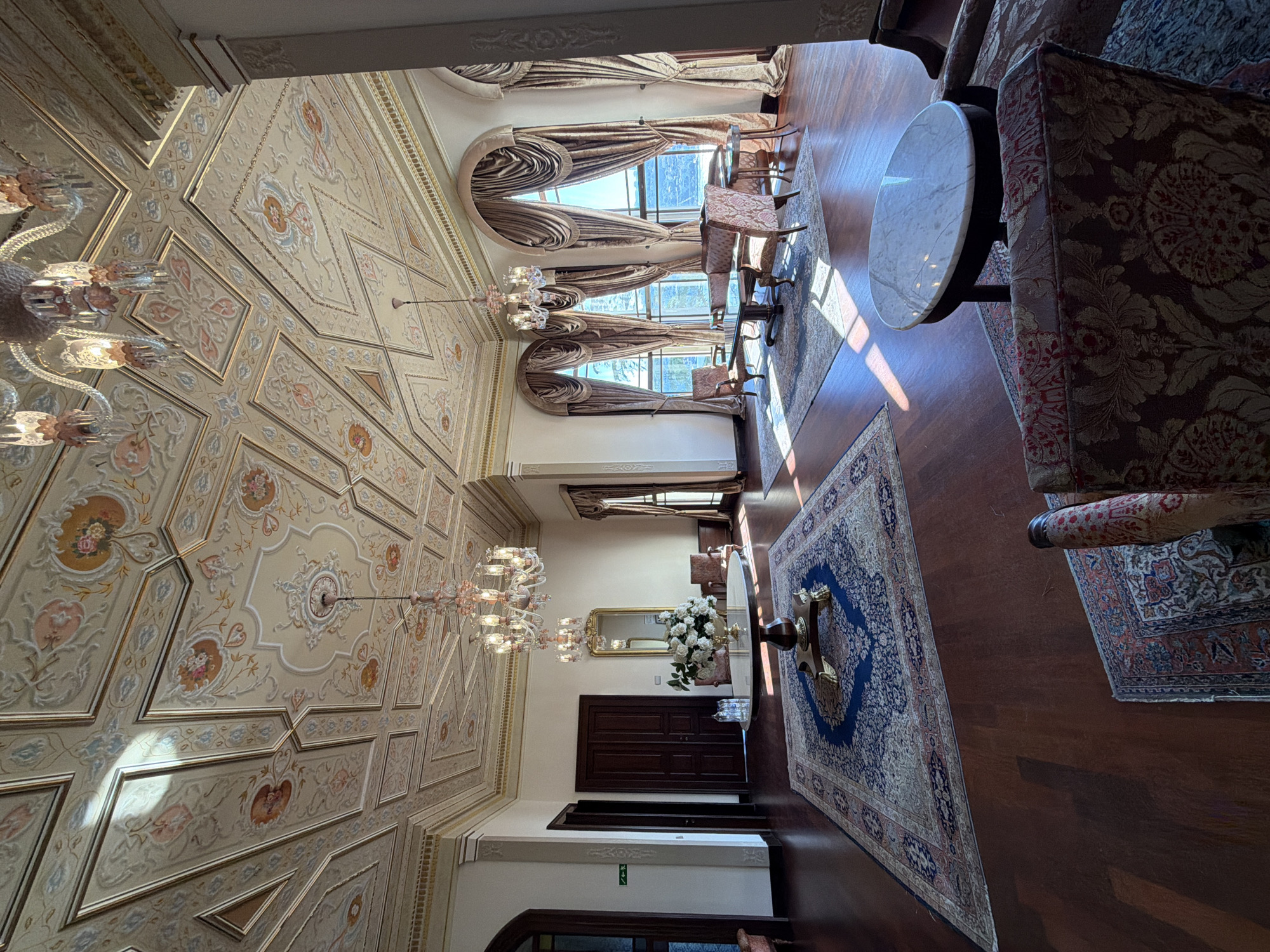 This is an interior shot of a grand, ornate room in the Beylerbeyi Palace in Üsküdar, Turkey. The room is decorated in a lavish style with a highly detailed, painted ceiling featuring floral motifs and geometric patterns. Two crystal chandeliers hang from the ceiling, casting a warm light. The floor is polished dark wood, and several large, intricately patterned Persian rugs are laid out.
In the center of the room, a large round table with a marble top stands on a decorative wooden base, adorned with a vase of white flowers. To the right, a seating area features two ornate chairs and a small table, positioned in front of tall, arched windows draped with heavy, golden curtains. Sunlight streams through these windows, illuminating parts of the room and casting dramatic shadows.
Further into the room, another seating area is visible with armchairs and sofas, suggesting a formal reception or drawing-room. The walls are painted in a pale color, accentuating the opulence of the ceiling and furnishings. Wooden doors are visible on the left side of the room. The overall impression is one of historical grandeur and luxurious elegance, typical of Ottoman palace architecture. There are no people present in the image.