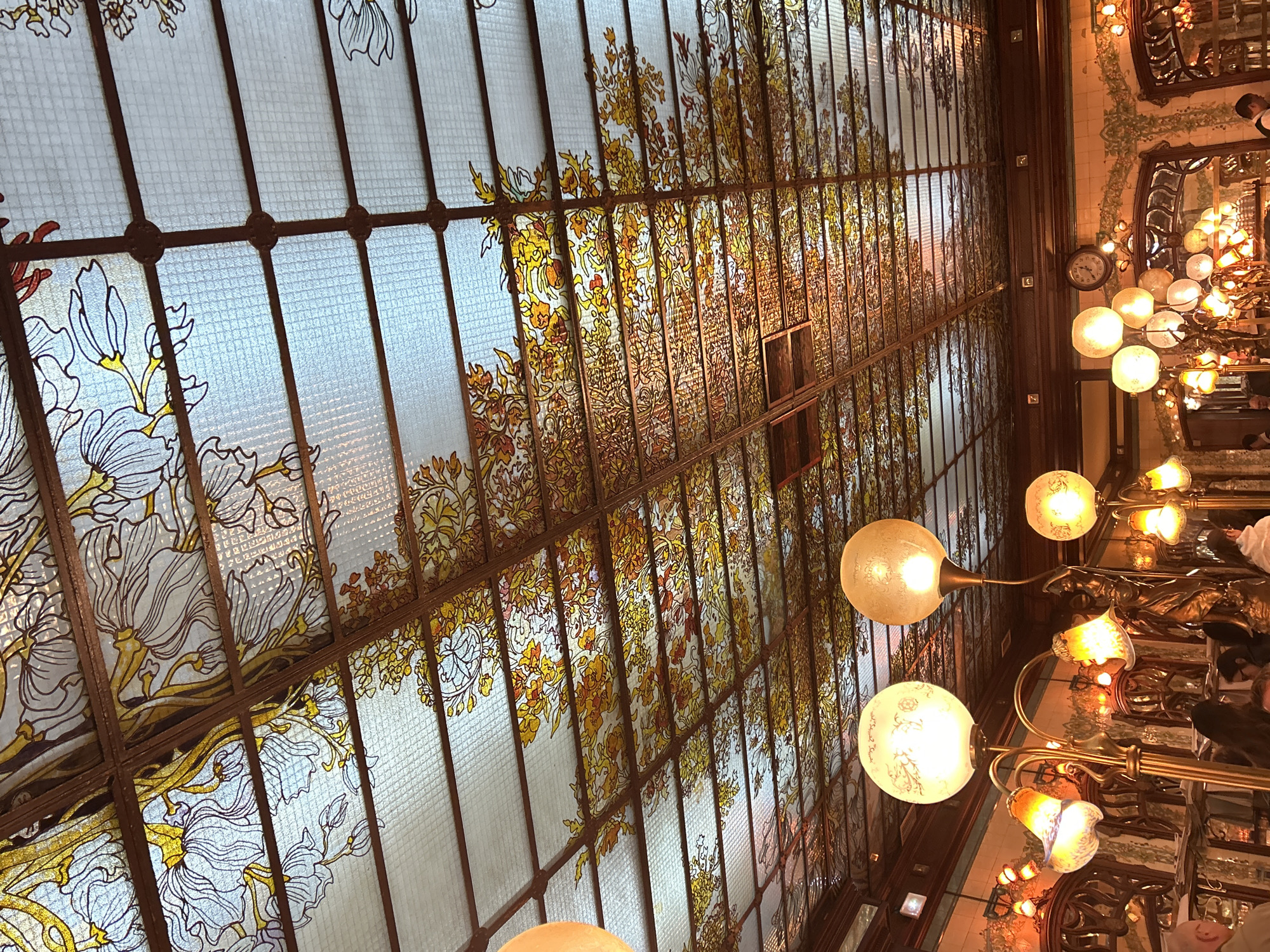 The image depicts an ornate, floral-patterned stained-glass ceiling, likely in an Art Nouveau style, above a restaurant interior. The ceiling consists of numerous rectangular glass panels separated by a grid of dark metal or wood supports. The glass is decorated with painted or stained floral motifs in shades of white, yellow, and brown.
Hanging below the ceiling are a series of elaborate light fixtures with spherical, frosted glass lamps and decorative metal arms. The fixtures provide ambient illumination.
In the background to the right, a portion of the restaurant interior is visible, including decorative mirrored walls, patterned tiles, a clock, and other light fixtures. People are seated at tables, their faces indistinct.
Based on the architectural style and the style of the restaurant, it is likely that the location is within Paris, France, as the city is known for its Art Nouveau architecture and historic restaurants.