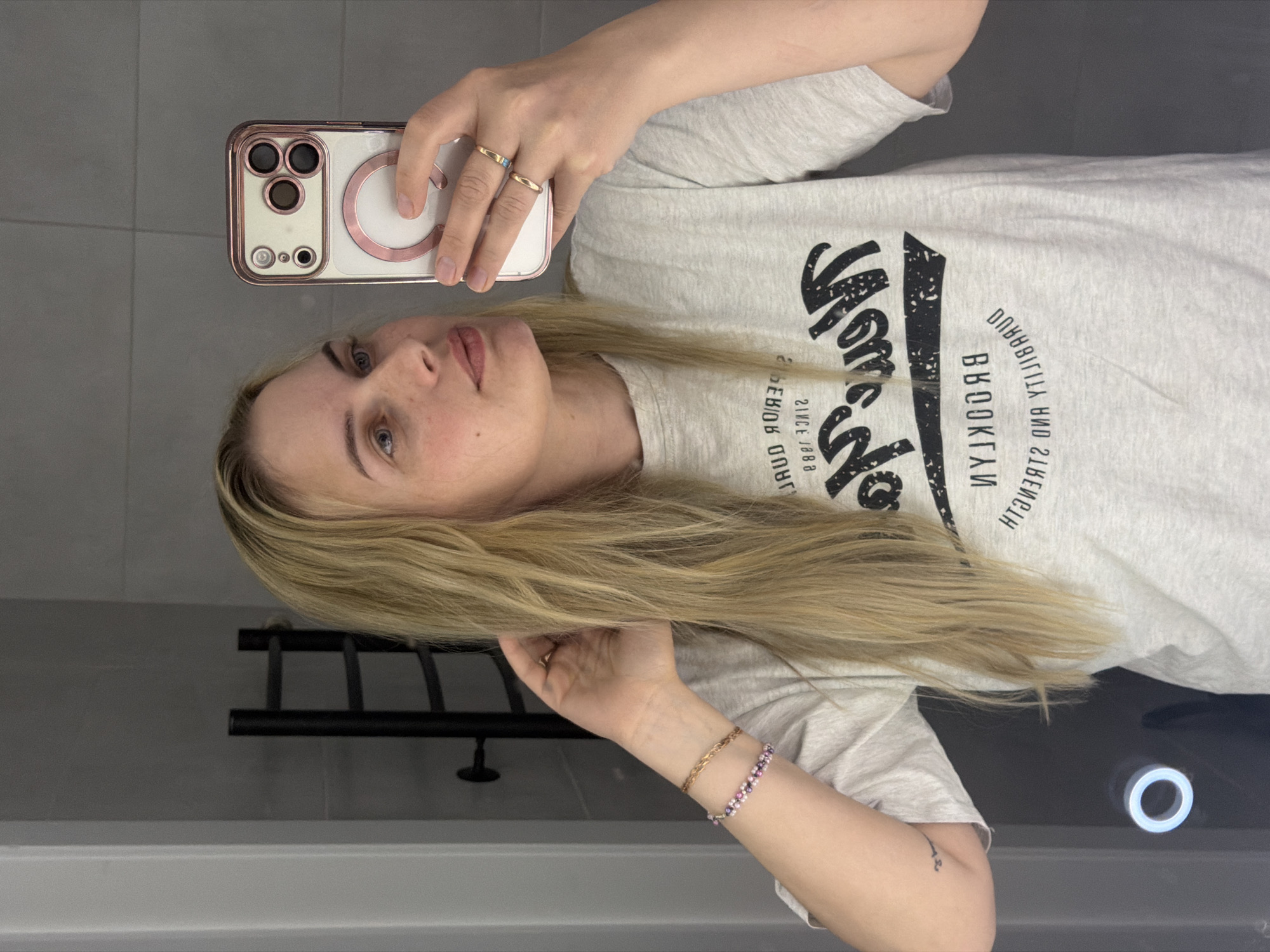 A fair-skinned woman with blonde hair is taking a selfie in what appears to be a bathroom. She is holding a white iPhone with a rose gold trim. She wears two rings on her left hand. She has a bracelet on her left wrist. She is wearing a light grey t-shirt with black lettering. The background shows grey tiles and a black towel rack. There is no information to suggest the image was taken in Chaikovsky, Russia.