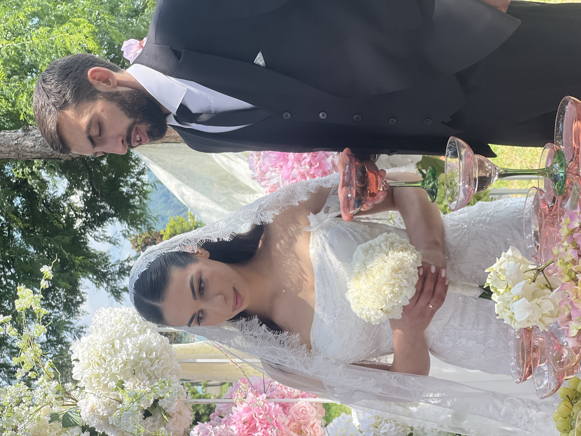 This image captures a wedding ceremony scene. Two people, a man and a woman, are the primary subjects. The man is wearing a black suit with a white shirt and black tie, a light pink flower pinned to his lapel. The woman is in a white wedding dress adorned with intricate lace and beading, and she wears a long lace veil. She is holding a small white bouquet.
They are standing outdoors, suggested by the lush green trees and foliage in the background, as well as the bright, natural lighting, indicating it's daytime. The ceremony appears to be set up with floral decorations, featuring an abundance of white and pink flowers, and a tiered stand holding pink-tinted glasses filled with a pink liquid, likely champagne.
The couple is engaged in the ritual of clinking glasses, a common element in wedding celebrations. The bride is looking down towards her bouquet and the glasses with a gentle, possibly content expression. The groom's face is partially obscured, but his gaze is also directed downwards. The overall mood seems joyous and celebratory, typical of a wedding day. There is no visible text in the image.