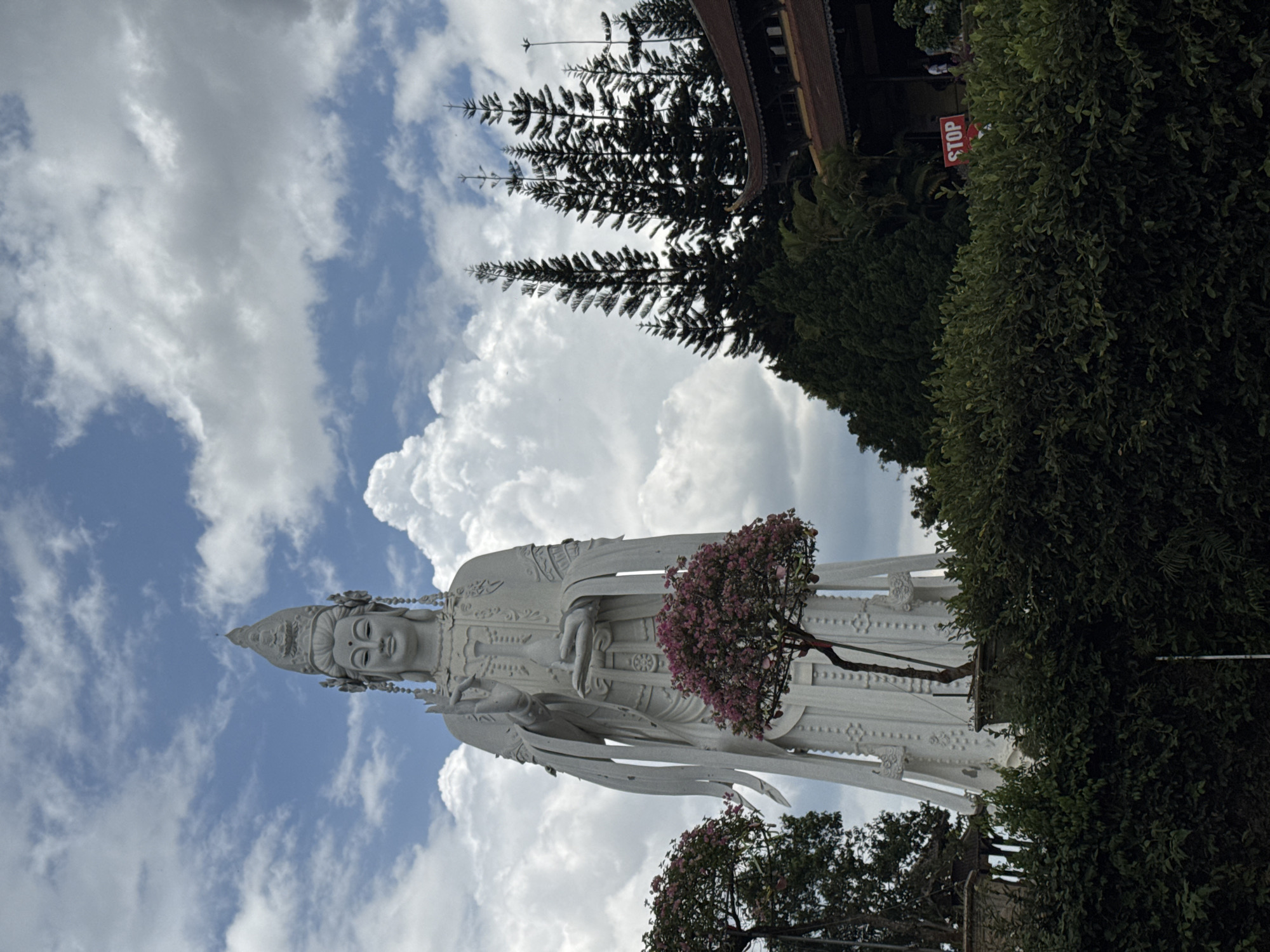 A low-angle, outdoor shot shows a large, white statue of a deity against a cloudy blue sky. The statue is slender and wears an ornate headdress. It holds an object in its hands. Pink flowers grow from a tree near the statue. To the right of the statue is a green bush and a tree. The top of the image includes the edge of a building and a "STOP" sign. The statue is of Guanyin, and is located in Linh An Pagoda, Nam Ban, Vietnam.