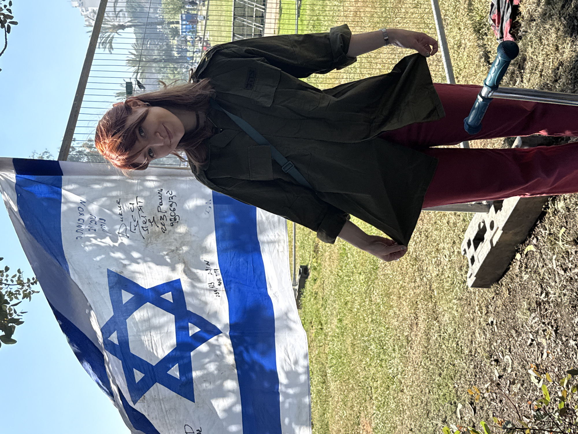 The image, captured at an oblique angle, shows a young woman standing outdoors in what appears to be Tel-Aviv, Israel, beside a large Israeli flag. The woman has long reddish-brown hair, illuminated by bright sunlight, and looks directly at the camera with a gentle smile. She wears an olive green military-style jacket or shirt, dark red pants, a dark choker necklace, and a silver bracelet on her left wrist, with some facial piercings visible. Her left hand is casually holding the hem of her jacket.
The prominent Israeli flag, white with two horizontal blue stripes and a central blue Star of David, is made of fabric and shows signs of use, with numerous handwritten messages, signatures, and dates scrawled across its surface in various languages, primarily Hebrew and Cyrillic script. Visible Hebrew phrases include "עם ישראל חי" (The people of Israel live), "יהי זכרם ברוך" (May their memory be blessed), and a date "23.10.23" (October 23, 2023), alongside other text. These inscriptions suggest the flag is part of a memorial, solidarity, or protest event.
Behind the woman and flag, a metal fence separates the immediate foreground from a grassy area with palm trees and distant buildings, indicating an urban outdoor setting. The ground is covered with grass and some dry earth. A blue scooter handle is partially visible to the right of the woman. The sky is clear and bright blue, suggesting a sunny day, likely morning or early afternoon, given the crisp lighting and shadows. The overall atmosphere conveys a sense of quiet solidarity or remembrance.