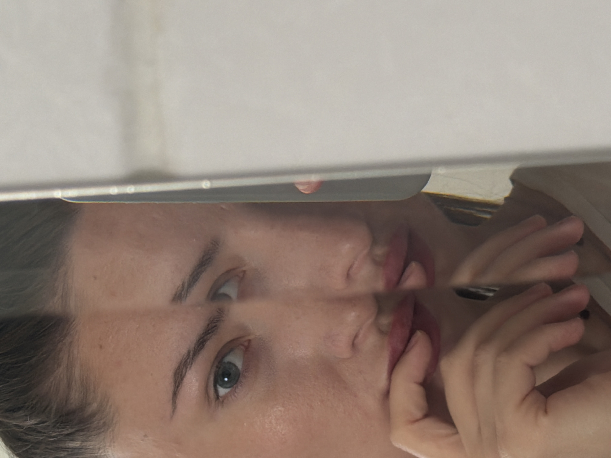 A close-up, distorted reflection shows a person's face and hands. The face is positioned in the left half of the image, with one eye clearly visible and a portion of the other. The skin appears fair, with visible pores. The eyebrows are thin and arched. The lips are painted a muted red and the person's fingers are placed near the mouth, with one finger touching the lower lip. The reflection creates a split image effect, with the right half of the face and body appearing slightly shifted. The background is a plain, light-colored surface, possibly a wall or screen, with a vertical line interrupting the reflection on the right side. There are no discernible elements in the image that would indicate a specific location in Sharm El Sheikh, Egypt.