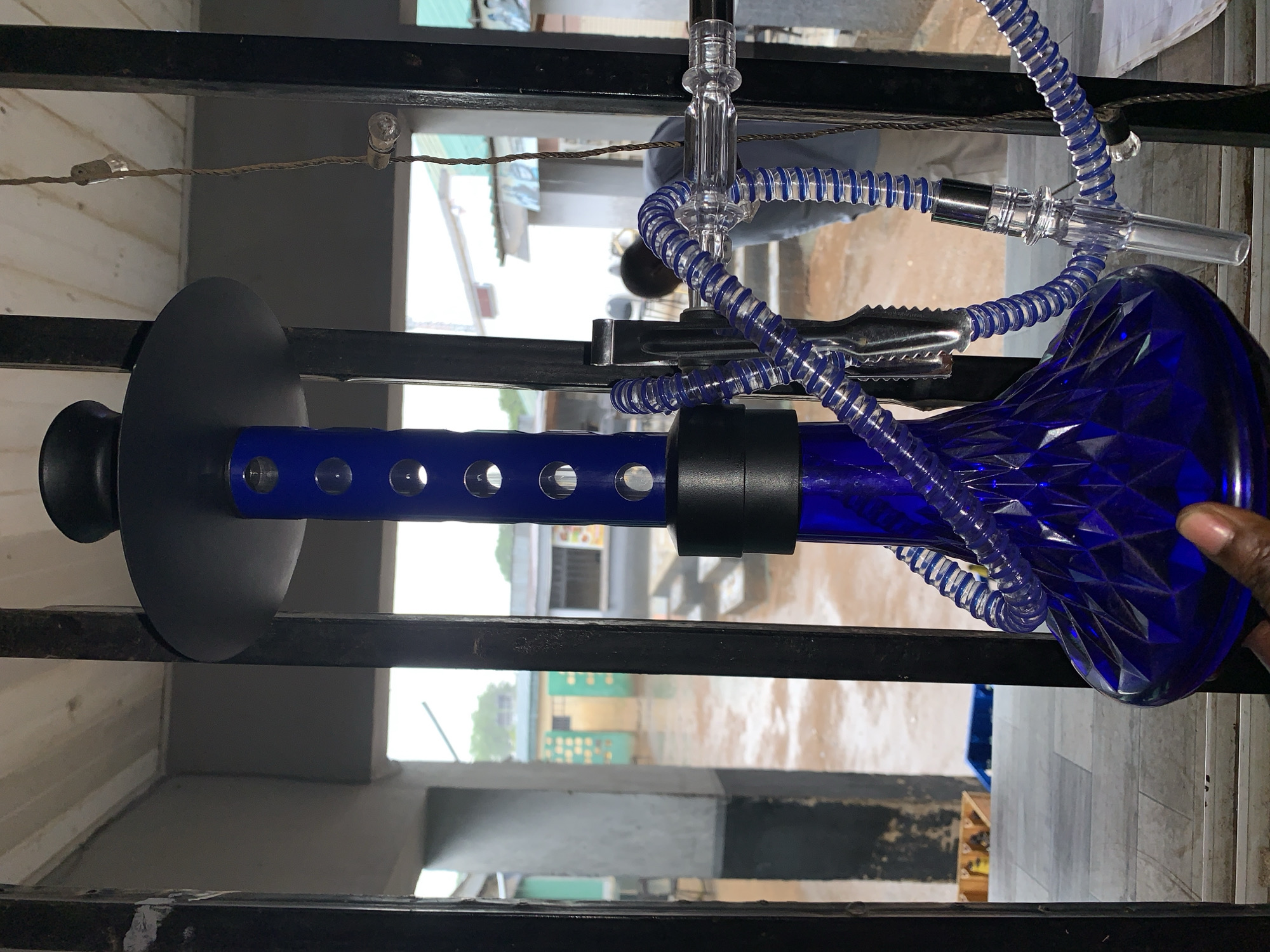 A close-up shot reveals a blue glass hookah set against the backdrop of Kabong, Nigeria. The hookah, featuring a blue vase with a textured pattern, is connected to a coiled blue hose. Black metal tongs are positioned near the hose. The hookah is held in place by a black metal rack. The environment visible through the rack consists of buildings in varying states of construction and roads. Light suggests daytime. An African hand is visible on the right side of the frame.