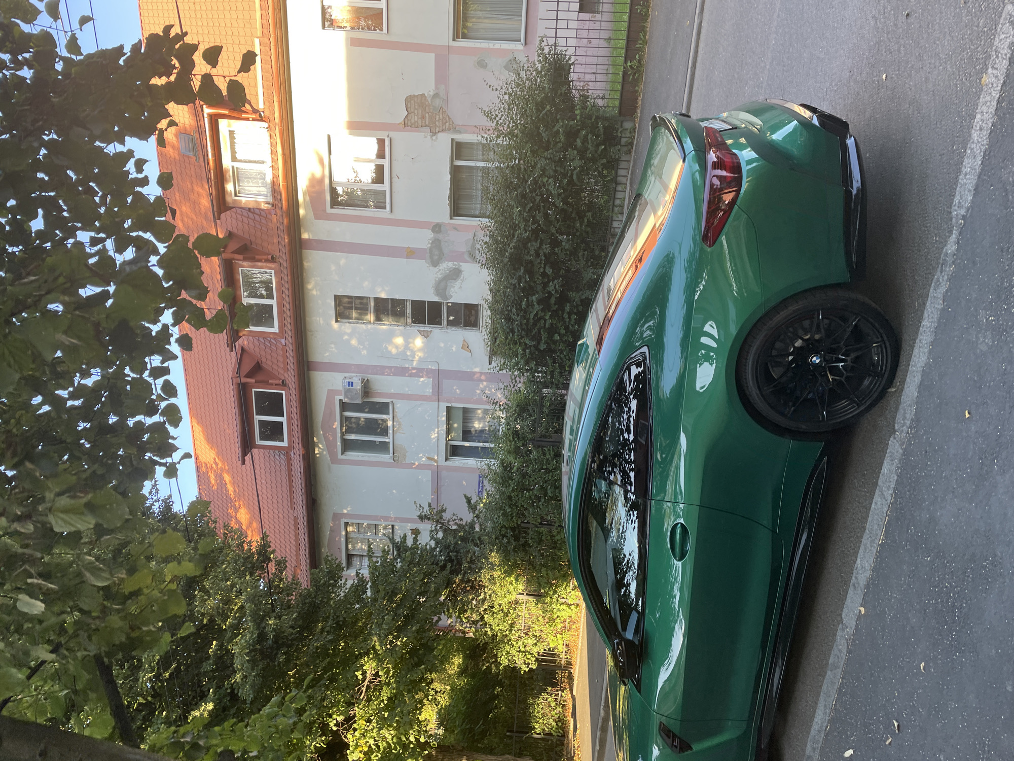 An eye-level, medium shot captures a bright green BMW M4 coupe parked on a paved city street, facing right. The car's sleek design and vibrant color are illuminated by the warm, golden light of the late afternoon sun, which casts long shadows and highlights reflections on the vehicle's surface.
Behind the car, a three-story building with a reddish-orange tiled roof and light pink and white facade stands. The building exhibits signs of wear, with sections of plaster peeling away to reveal brickwork. Several windows, some with curtains and others with air conditioning units, are visible on its exterior. The building's facade is decorated with thin pink vertical stripes.
To the left of the building, a lush green tree with dense foliage overhangs the scene, its leaves catching the sunlight. A black metal fence runs along the property line between the building and the street. In front of the building, a row of well-maintained green bushes partially obscures the lower story.
The street is paved with asphalt, showing some minor wear and tear. A concrete curb separates the street from a narrow strip of sidewalk or verge where fallen leaves are scattered. The sky above is a clear, light blue, indicating fair weather. The overall impression is a quiet urban setting bathed in the soft light of a summer evening. The scene is devoid of any people.