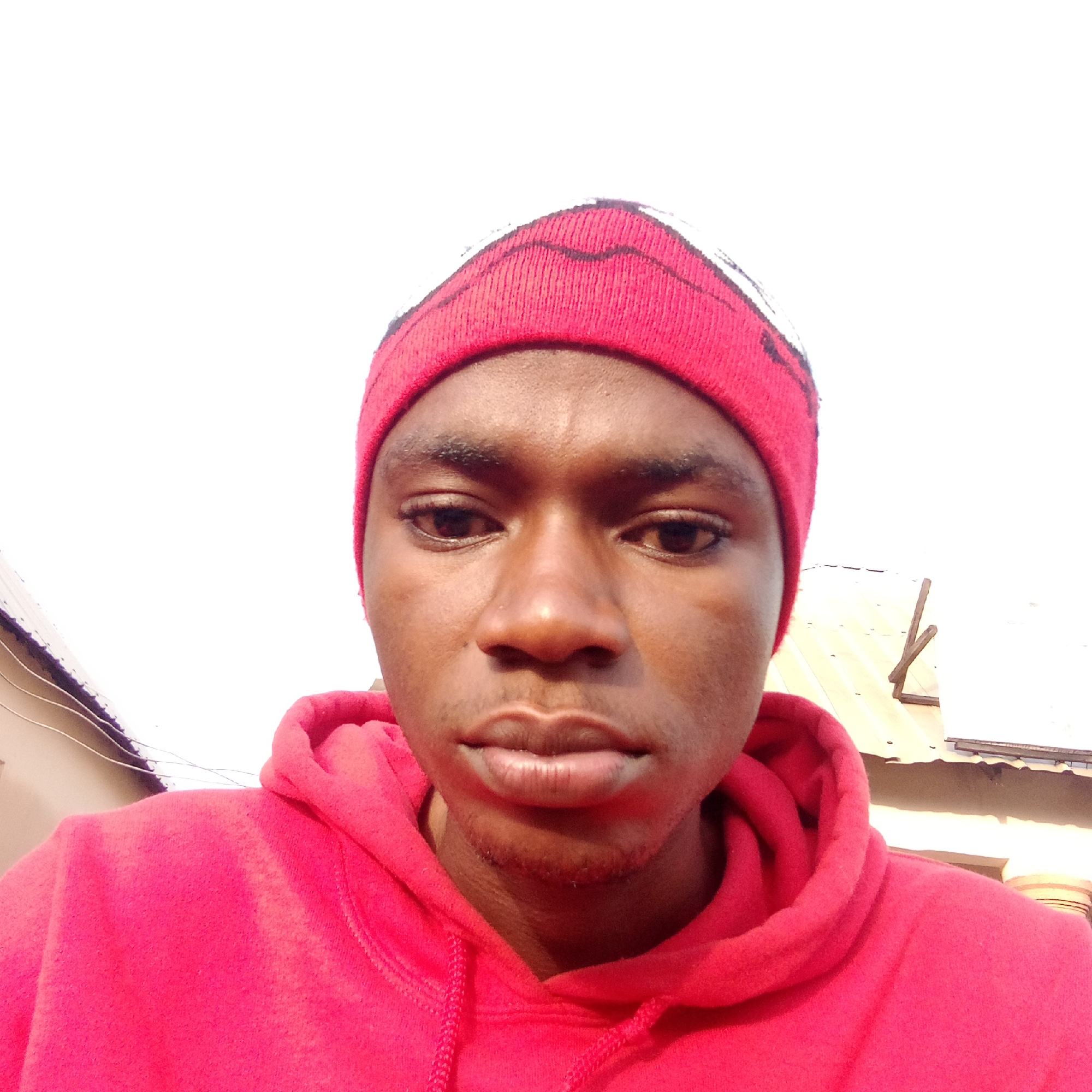 A close-up, low-angle shot shows a young, dark-skinned man wearing a red hooded sweatshirt and a red knit cap with a white accent. The man is the primary subject, and he is looking directly at the camera with a neutral expression. The background is indistinct, but it appears to be a residential area with a corrugated metal roof visible on one side and a building with a wire extending from it on the other. The man's features are clearly visible, including his eyes, nose, and lips. The setting is consistent with an outdoor location during the day. Based on the prompt, the location is identified as Mararaba, Nigeria.