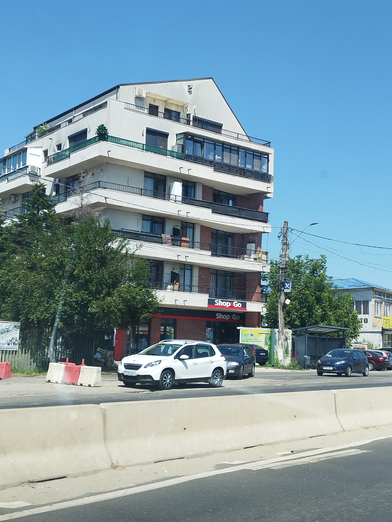 A modern, multi-story apartment building with balconies and large windows stands on the left side of the frame, adjacent to a commercial establishment with "Shop Go" signage. In front of the building, a white Peugeot 208 hatchback is parked. Several other vehicles are visible on the road, including a dark blue hatchback. The scene is set on a sunny day with a clear blue sky. The foreground features a concrete traffic barrier. The location is Bragadiru, Romania.