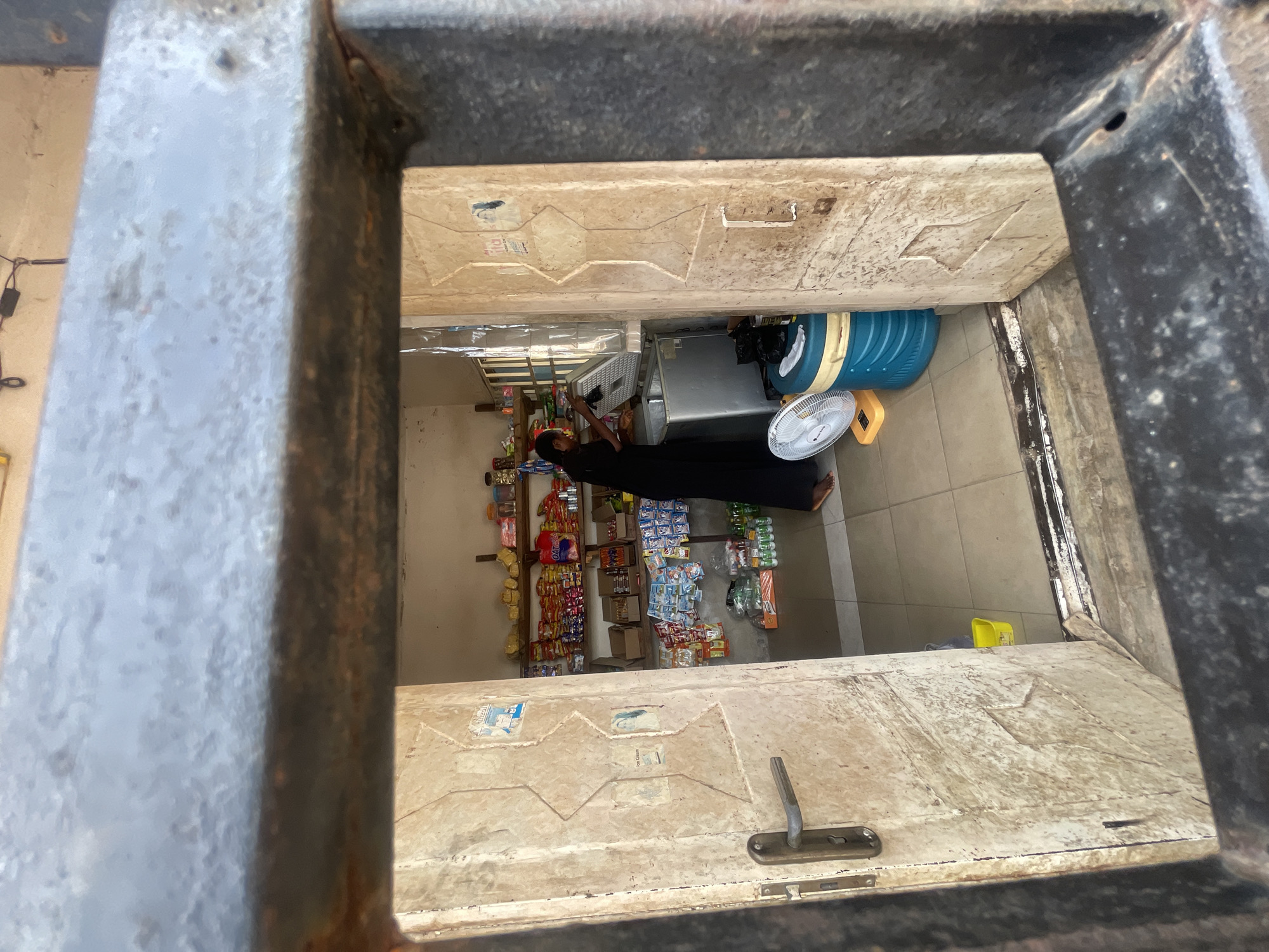 View from above, through a square window of a shop in Lokoja, Nigeria. A person in a long black dress stands inside. Shelves stocked with snacks line one wall. A cooler, a fan, and a large blue water jug are also visible. The walls are painted white. The window frame is metal, coated with gray paint, showing signs of rust and wear. A white painted door with diamond designs is partially opened in the window.