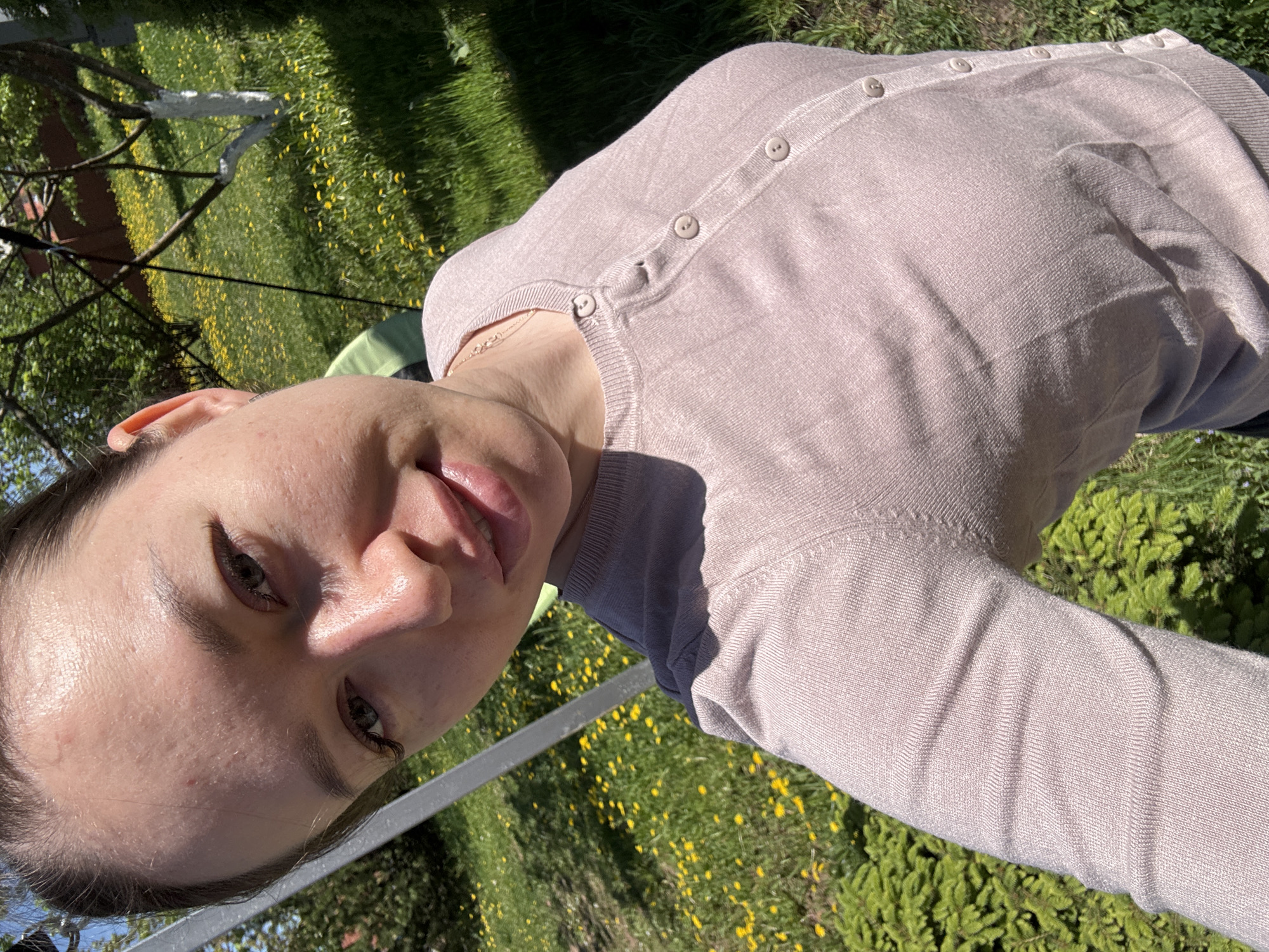 A woman is posing for a selfie outdoors in Ферма, Russia. She is wearing a light pink cardigan and a delicate gold necklace. The background shows a lawn filled with yellow wildflowers, likely dandelions. A metal structure, possibly part of a chair or other garden furniture, is partially visible on the left side of the frame. The lighting suggests a sunny day.