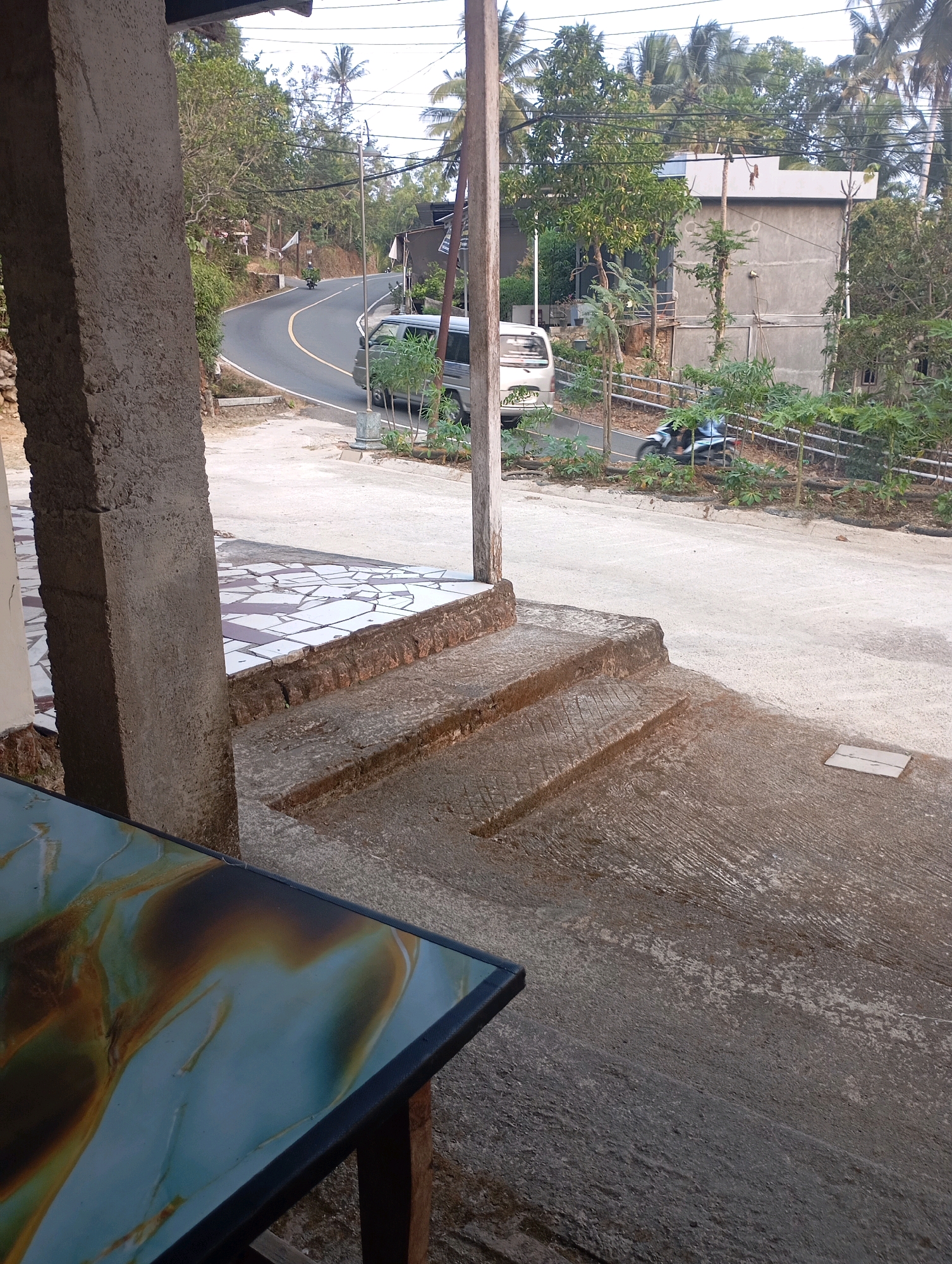 The image depicts a scene in Ketepung, Indonesia, likely a residential or rural area. In the foreground, a rough concrete pillar stands to the left, partially obscuring a table with a patterned surface. Adjacent to the pillar are a series of outdoor steps, constructed from concrete and bordered by a tiled platform. The steps appear weathered and somewhat worn.
Beyond the steps, a paved road curves into the distance. A white van is parked on the side of the road, and a motorcycle is being ridden away from the viewer. Lush green vegetation and trees line the sides of the road, with a few palm trees visible against the sky. In the background, simple buildings with concrete walls and tiled roofs can be seen amongst the trees. The sky is overcast, suggesting either early morning or late afternoon lighting. The overall impression is one of a quiet, everyday scene in a tropical setting.