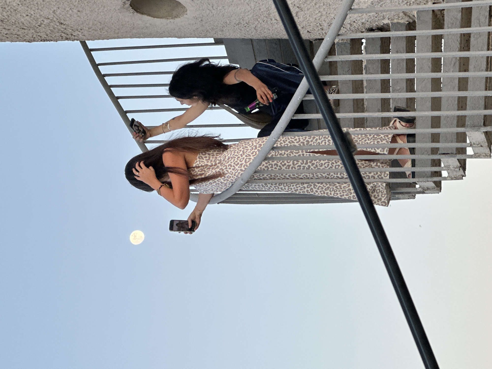 Two women are on a metal staircase. The woman in the foreground is wearing a leopard-print dress and holding a cell phone, presumably taking a photo of the full moon in the sky. The other woman is kneeling behind her on the staircase, also holding a cell phone, likely taking a picture or video. The location is Tashkent, Uzbekistan.