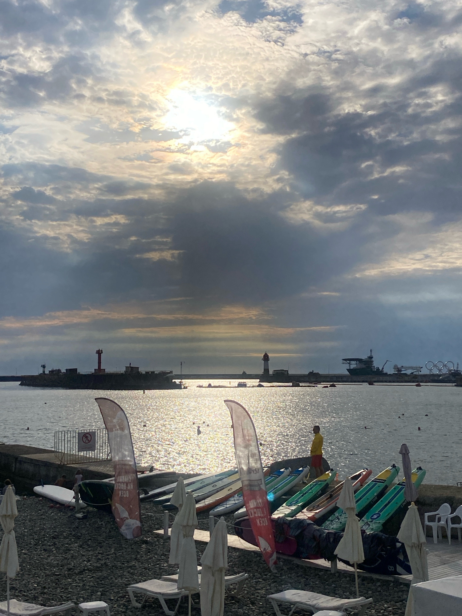 The image depicts a waterfront scene, likely in Sochi, Russia, given the Olympic rings visible in the distance. The sky is overcast with the sun breaking through the clouds, casting a bright glare on the water. In the foreground, a pebbled beach is dotted with lounge chairs and closed umbrellas. Several paddleboards are neatly arranged along the edge of a paved area, some leaning against a railing. A person wearing a yellow shirt and red shorts stands near the paddleboards, observing the water. In the background, a harbor is visible, featuring lighthouses, a ship, and the iconic Olympic rings structure. The water is choppy and reflects the sunlight. A fence with a warning sign is also present near the water's edge.