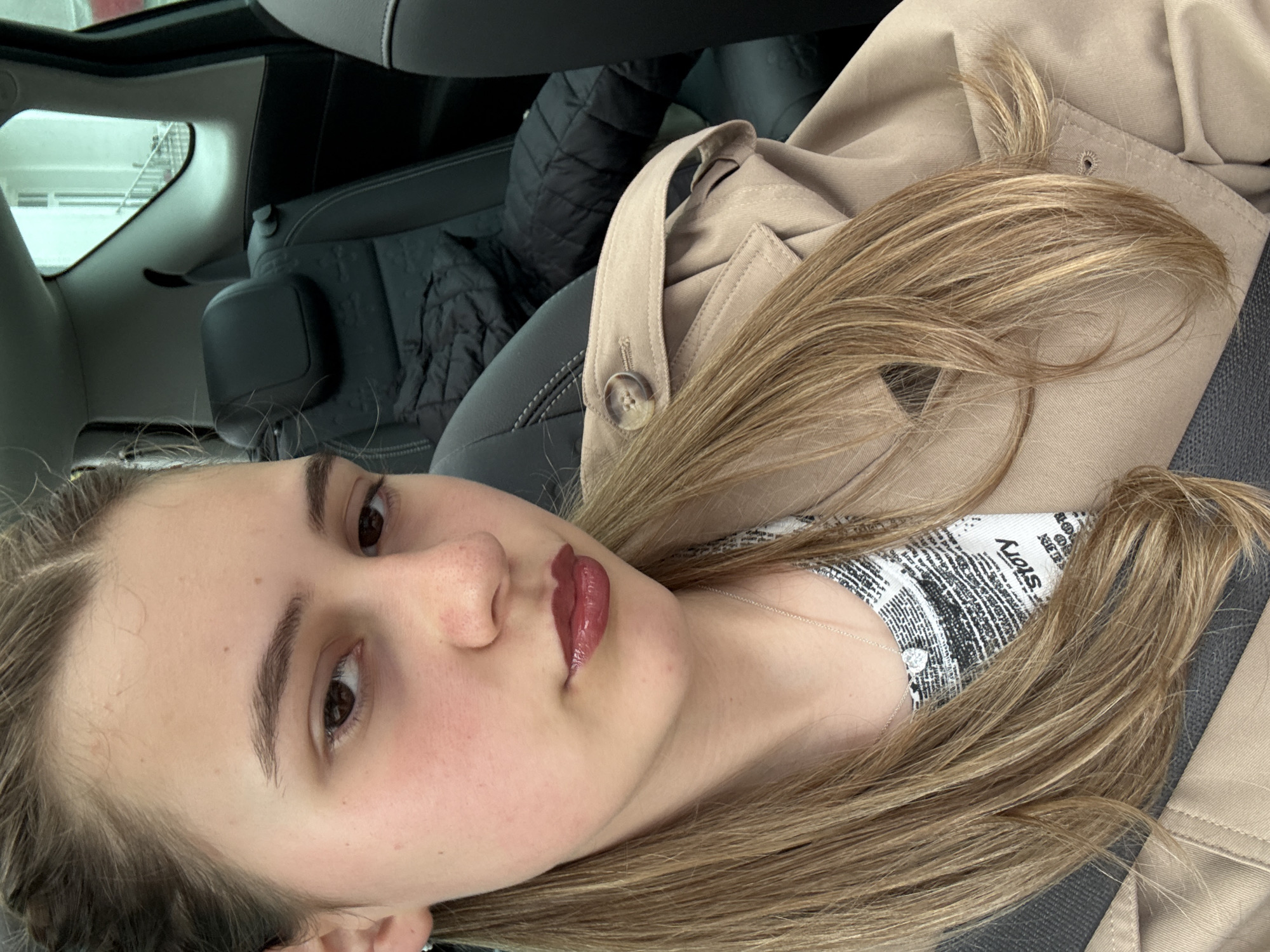 A light-skinned woman with light brown hair is seated in a vehicle. She has brown eyes, and her lips are painted with a glossy maroon-brown lipstick. She's wearing a light beige jacket, and beneath it, a patterned top with newspaper print is visible around the neckline. A delicate silver necklace with a small pendant sits on her chest. The interior of the car includes black leather seats. Based on the available image, the specific location in Panagyurishte, Bulgaria cannot be determined.